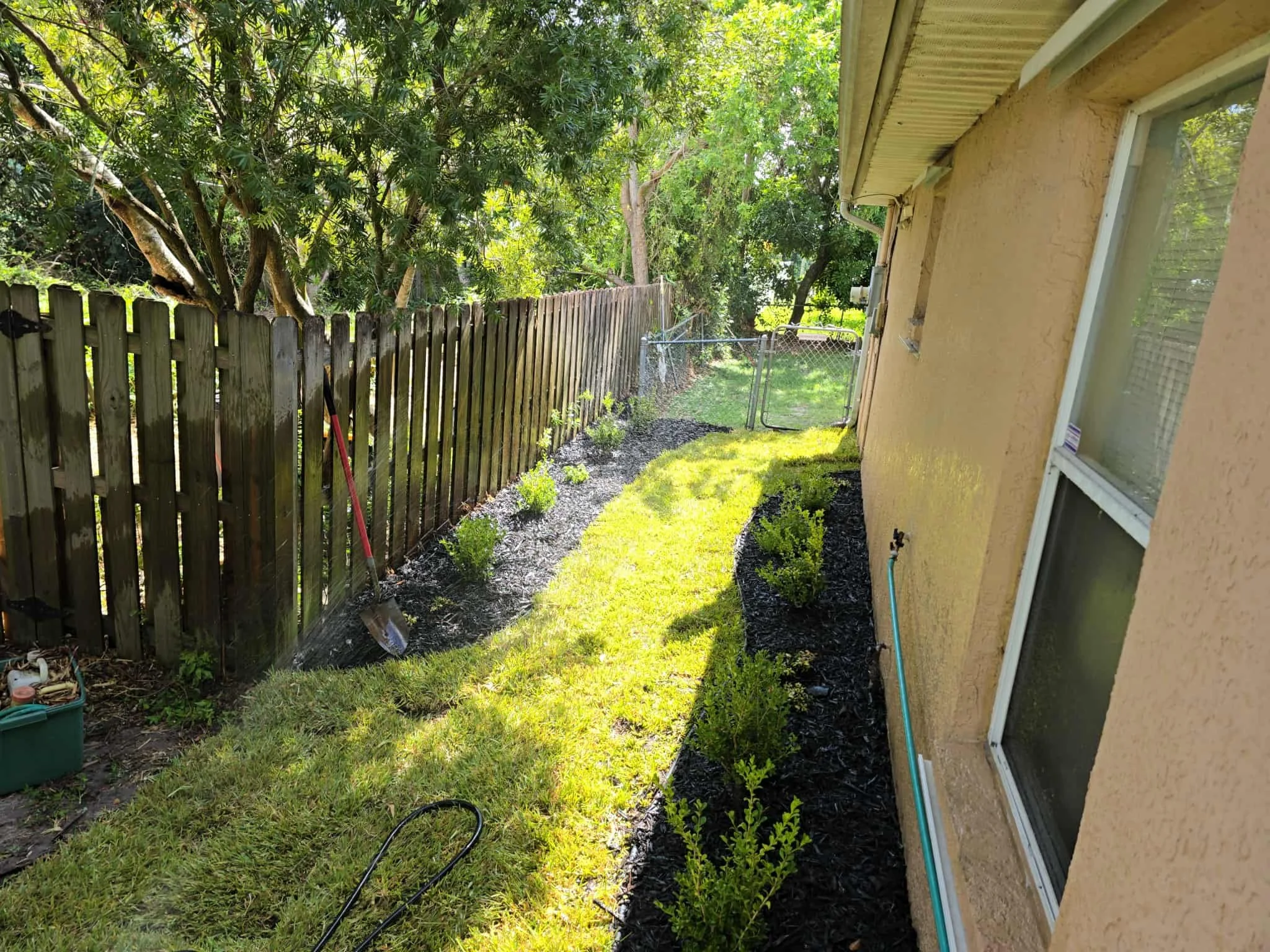 A backyard with a yellow house wall, windows, and a garden. There is a wooden fence, green grass, bushes, and trees providing shade. A garden bed with small plants is along the house, and garden tools are visible.