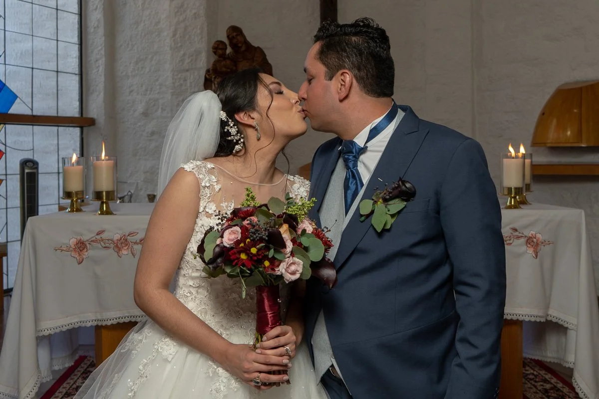 Bride and groom kissing during their wedding ceremony, bride holding a bouquet of flowers, candles on table behind them.