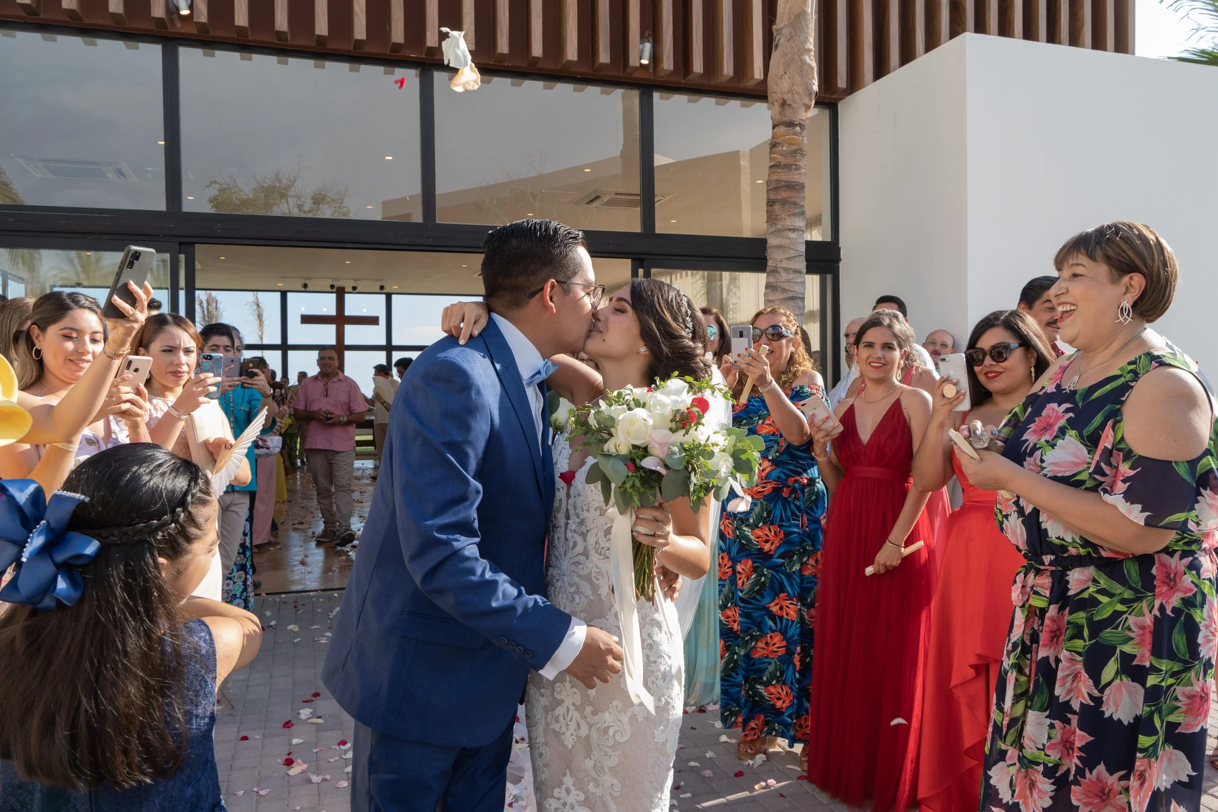 Pareja de recién casados besándose frente a amigos y familiares en una boda al aire libre, con muchas personas sonriendo y tomando fotos.