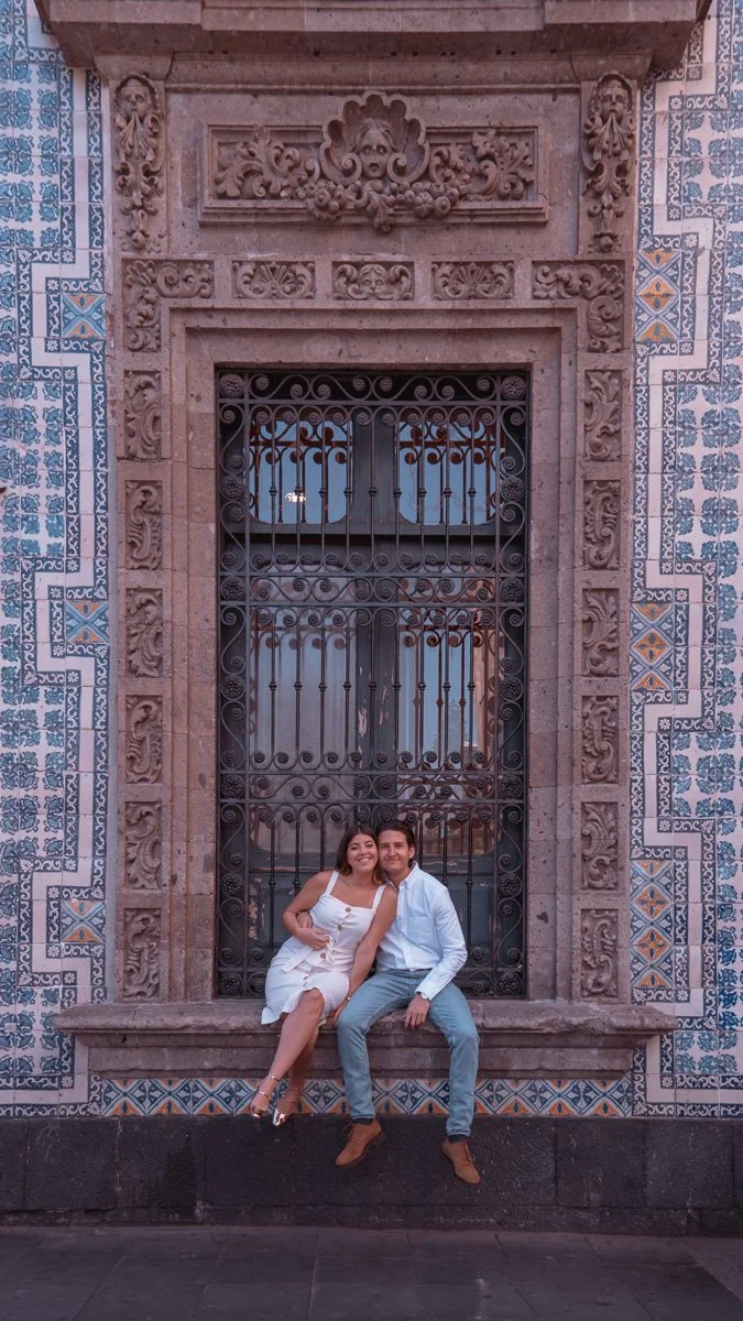 A smiling couple sitting on a stone ledge in front of an ornate, wrought iron gate set into a historic stone building with decorative tilework.