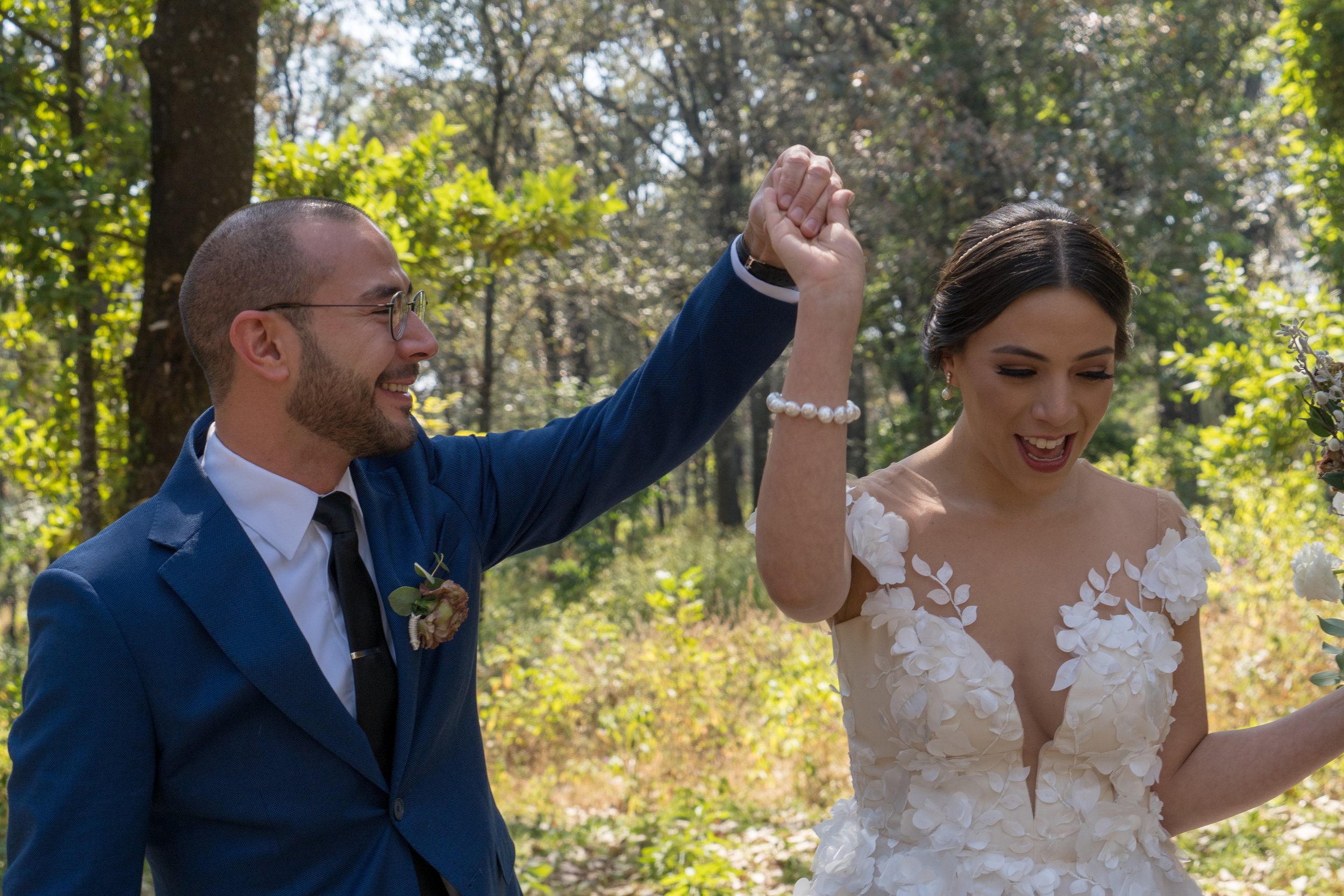 Pareja de recién casados en una ceremonia al aire libre en el bosque, la mujer lleva un vestido de novia con flores y el hombre un traje azul con corbata. Están celebrando y levantándose las manos.