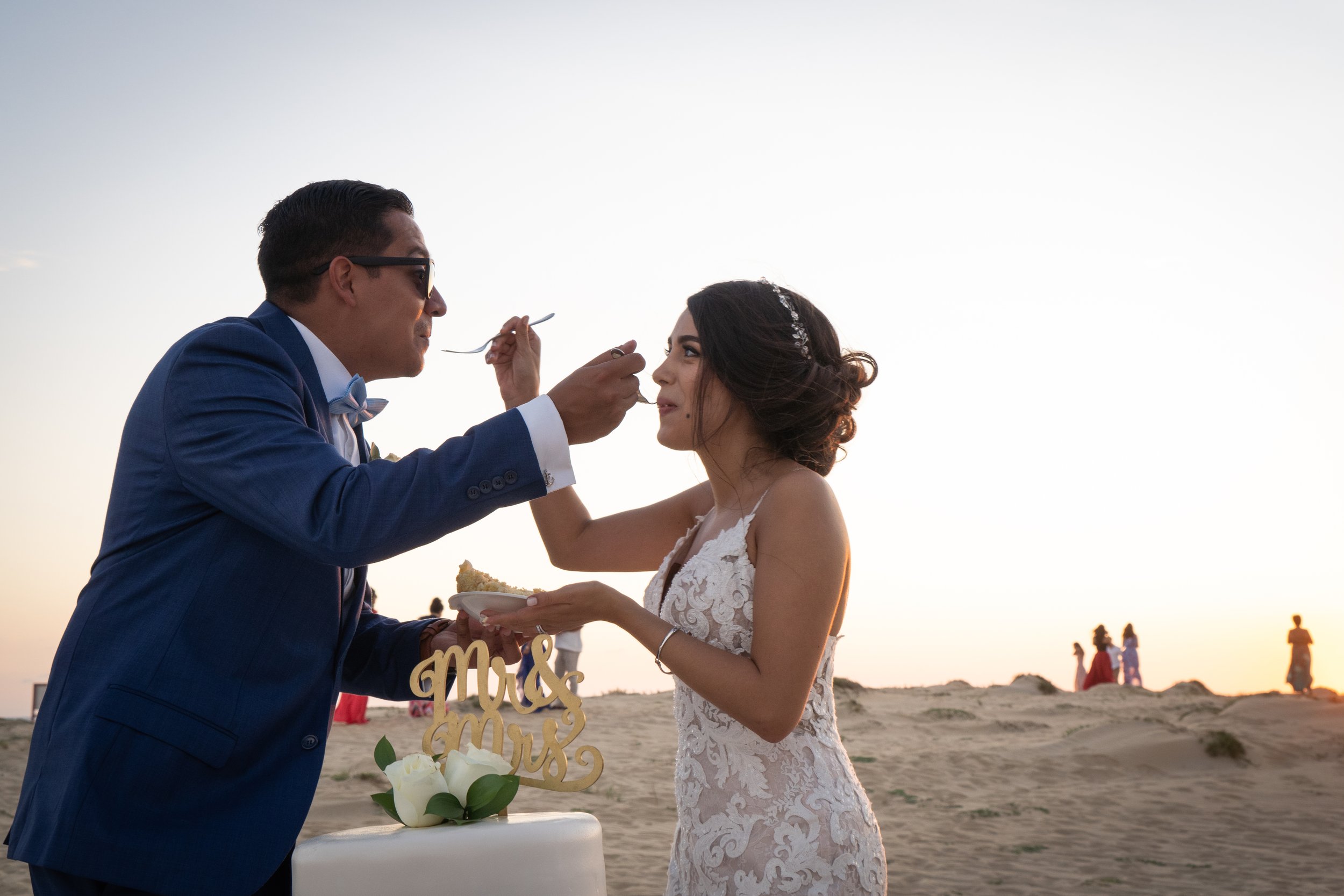 Pareja de recién casados en la playa, la novia recibe comida de la boda, al fondo se ven otras personas y el atardecer.