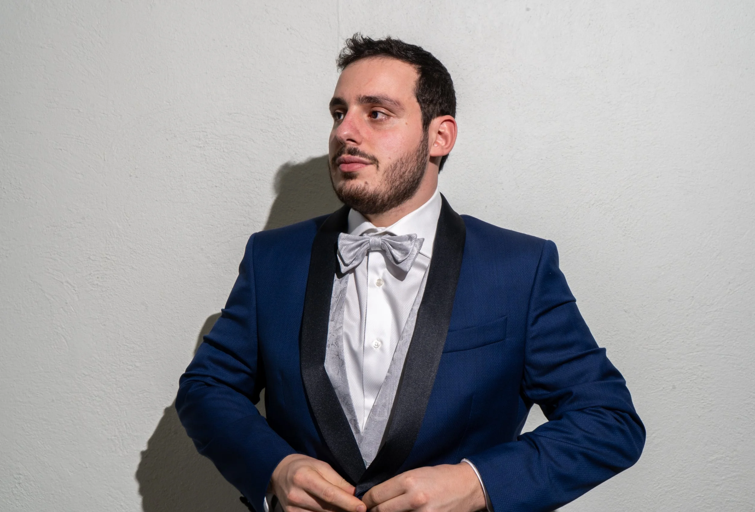 Un hombre con barba, cabello oscuro, vestido con esmoquin azul y una pajarita gris, ajustándose la chaqueta