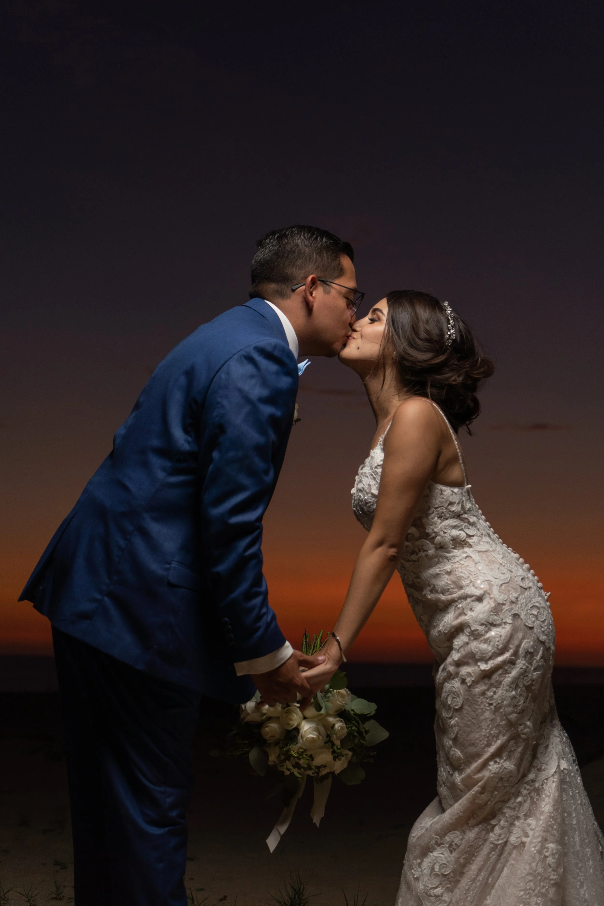 Una pareja de recién casados se besa en la playa al atardecer, la mujer con vestido de bodas y el hombre con traje azul, sosteniendo un ramo de flores blancas.