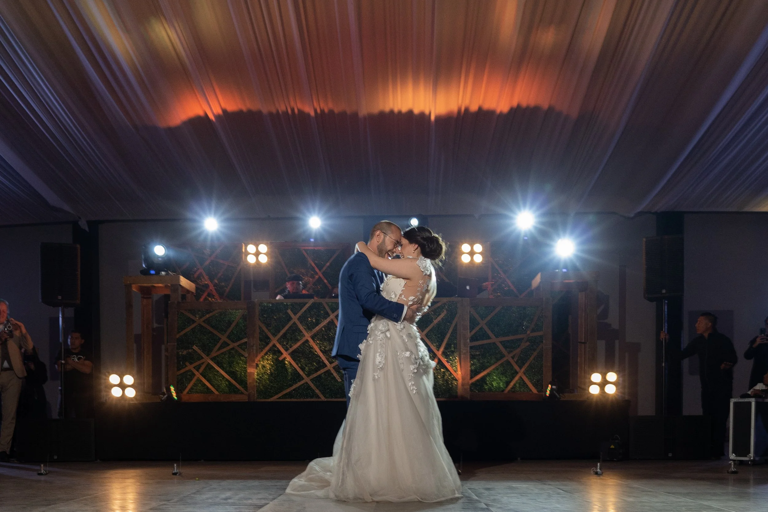 Una pareja bailando en una boda, en una pista iluminada con luces brillantes y un DJ en el fondo.
