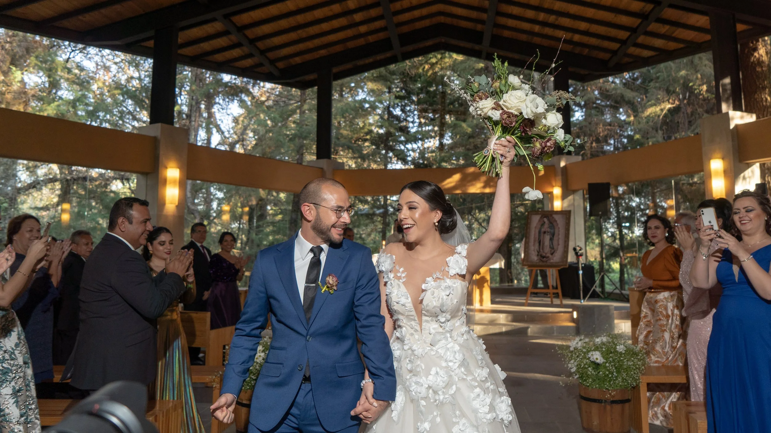Una pareja de recién casados en una ceremonia en un entorno natural, la novia sostiene un ramo de flores en alto, rodeados de amigos y familiares que los felicitan y toman fotos.