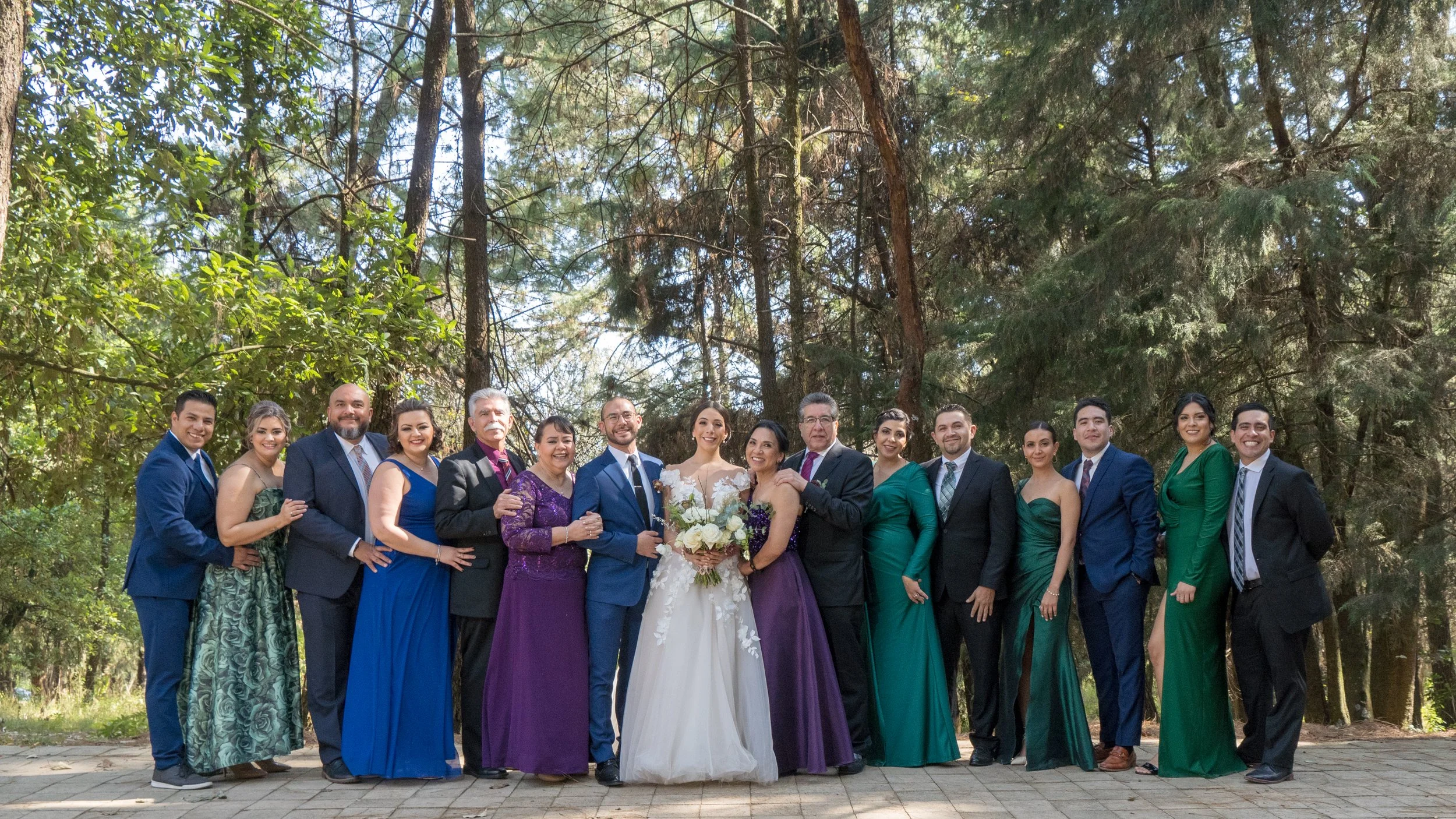 Grupo de personas en una boda, posando en un bosque, con el conductor de la novia en el centro y todos sonriendo.