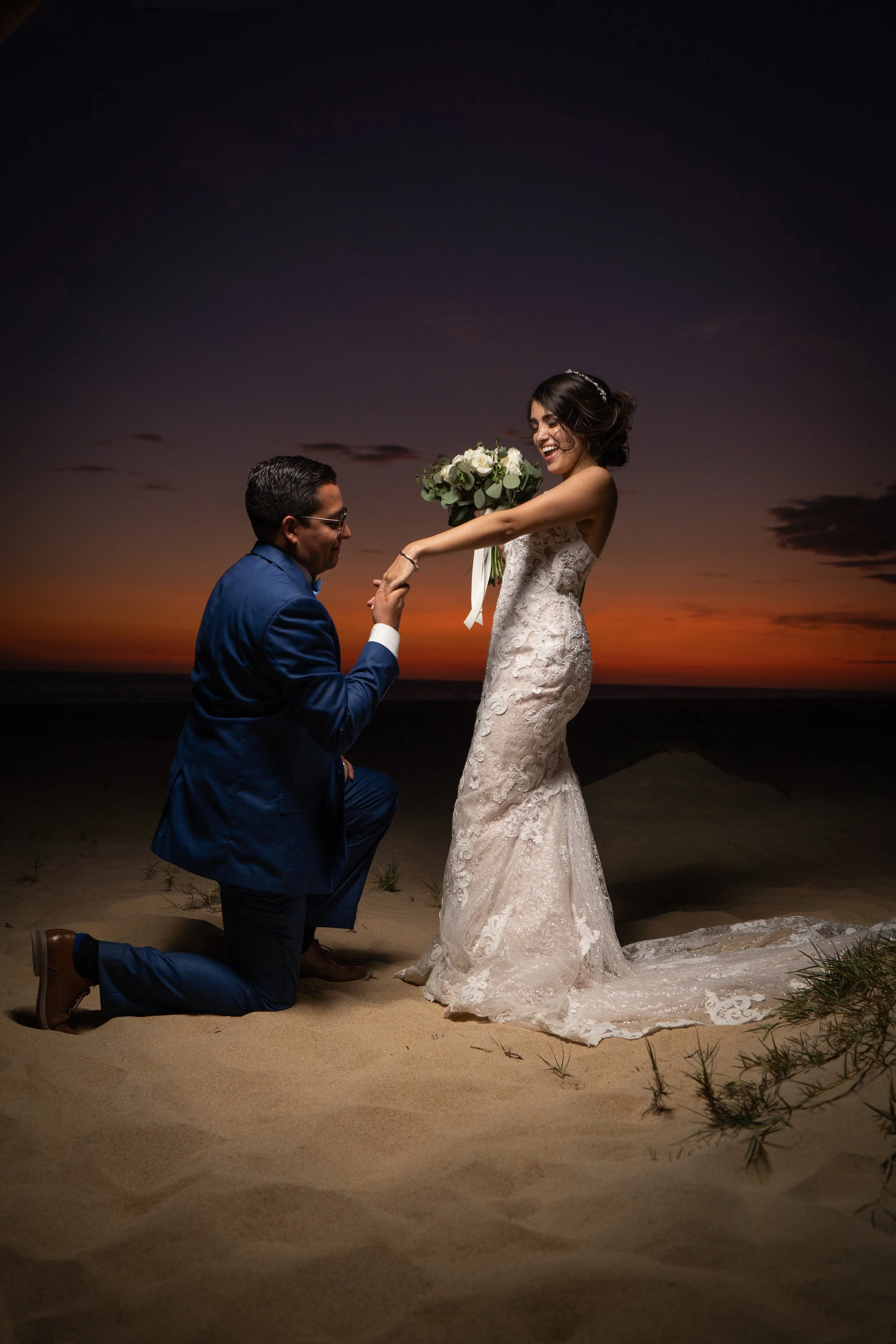 Pareja de novios en la playa durante el atardecer, la novia sostiene un ramo de flores y el novio la está ayudando a levantarse, ambos sonrientes y felices.