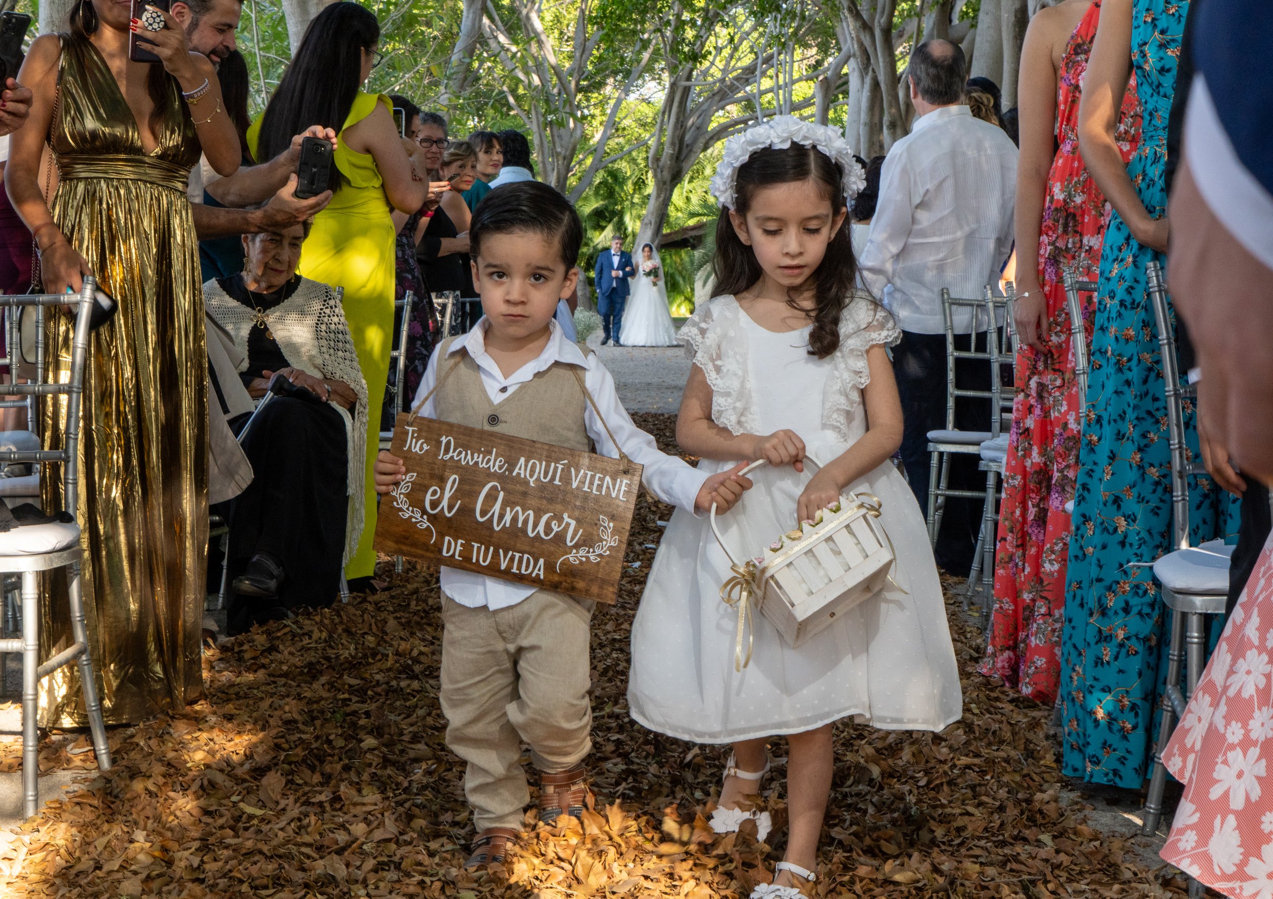 Niños participando en una ceremonia de boda. Un niño sostiene un cartel que dice 'Tío Davide, AQUÍ VIENE el Amor DE TU VIDA' y una niña lleva una cesta pequeña con flores.