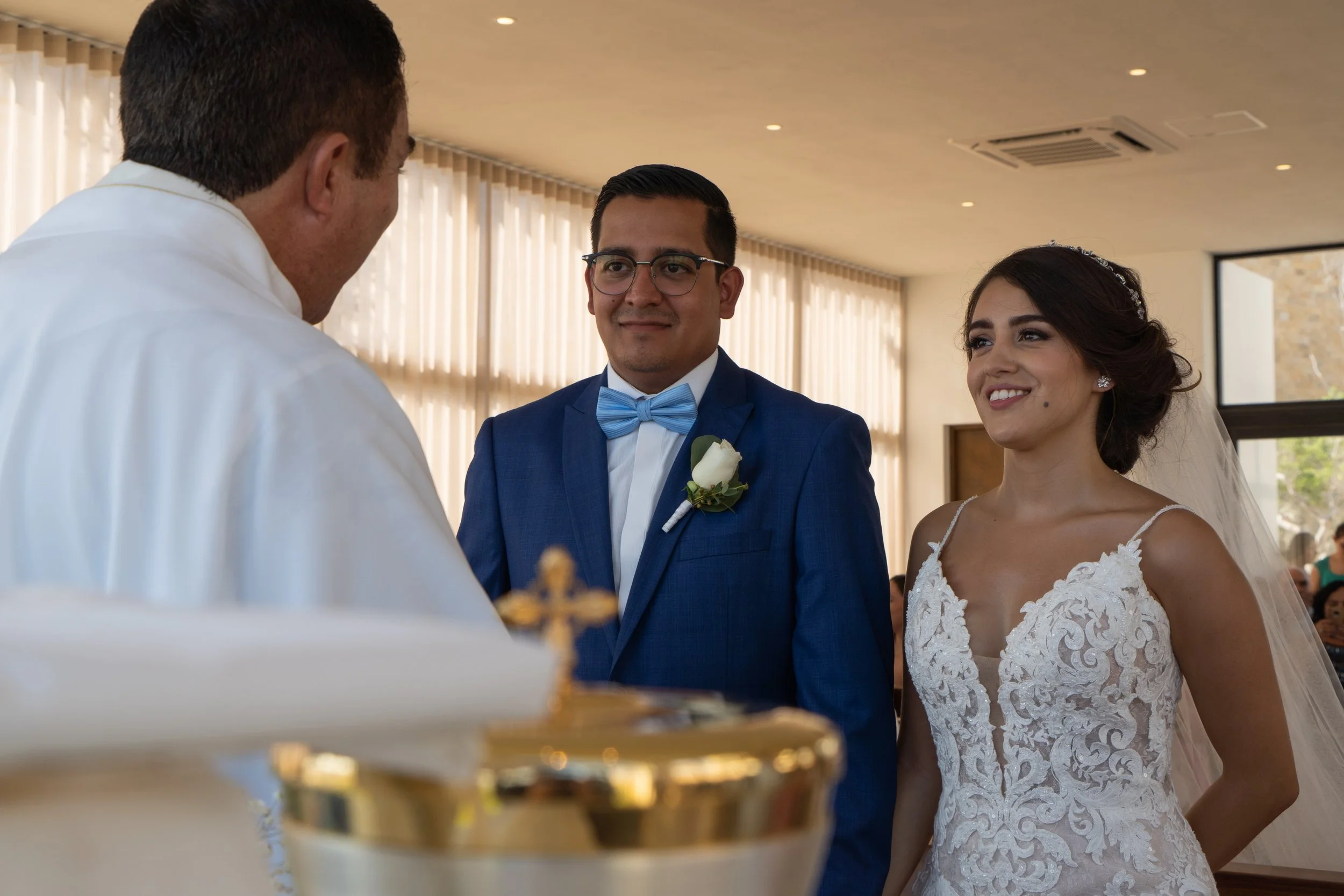 Una pareja de novios en su boda, la novia lleva vestido blanco con encaje y velo, el novio viste traje azul con pajarita, están en una ceremonia religiosa con un sacerdote frente a ellos.
