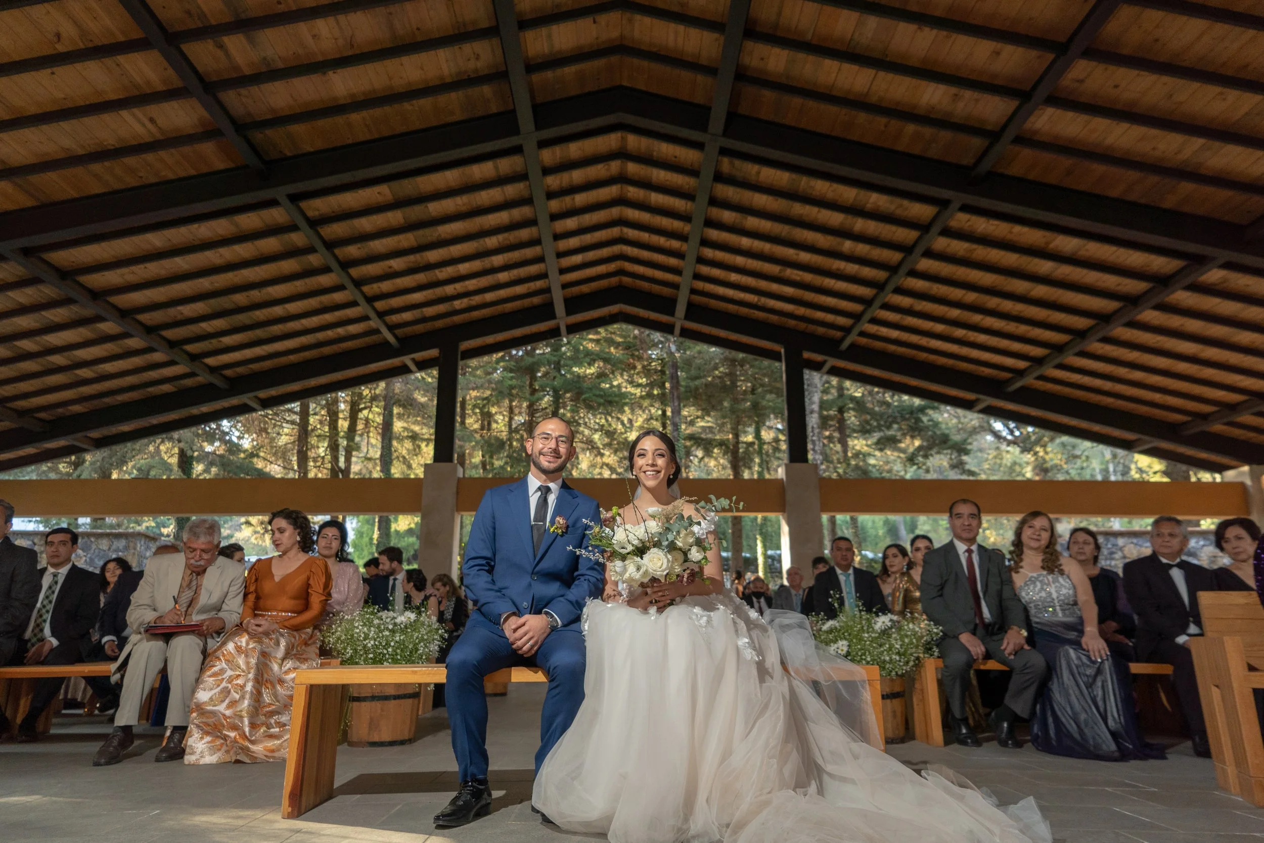Pareja de novios en la ceremonia, sentados en un banco, rodeados de invitados en un espacio abierto y rodeado de árboles.