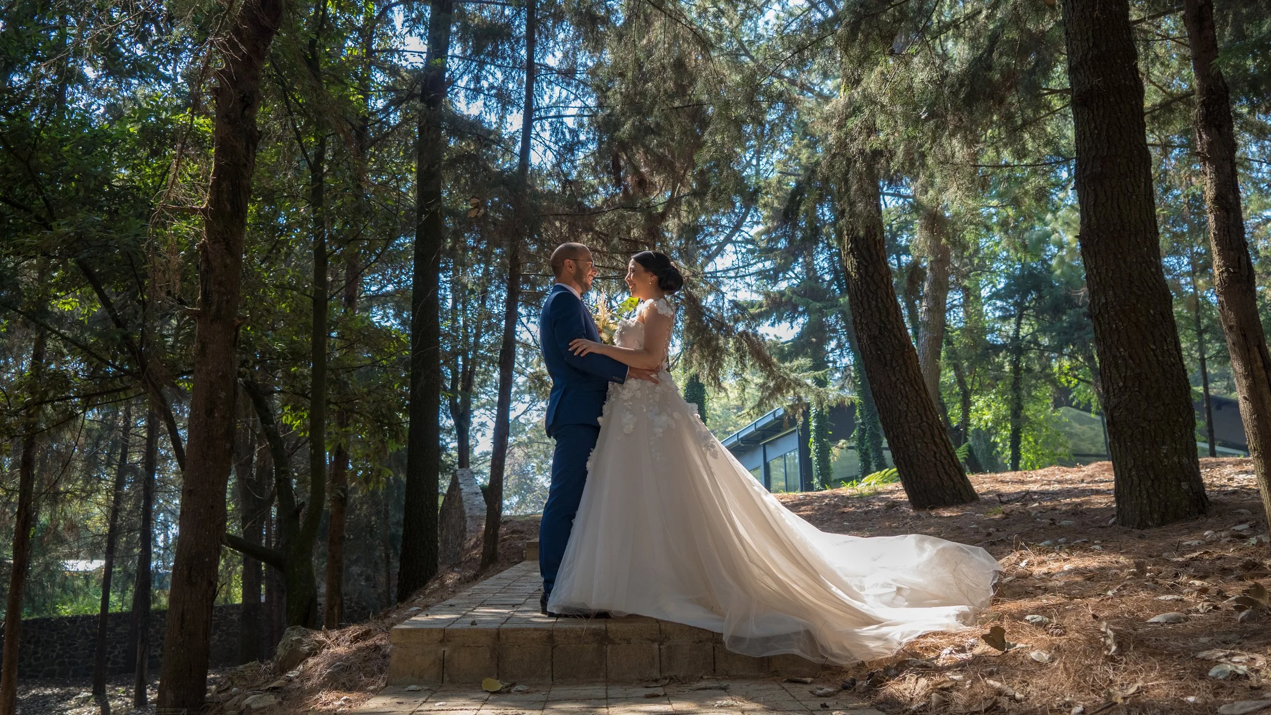 Una pareja de novios se encuentra en un bosque, con árboles altos y luz solar brillante, en una ceremonia de boda al aire libre. La novia viste un vestido de novia largo y el novio un traje azul.