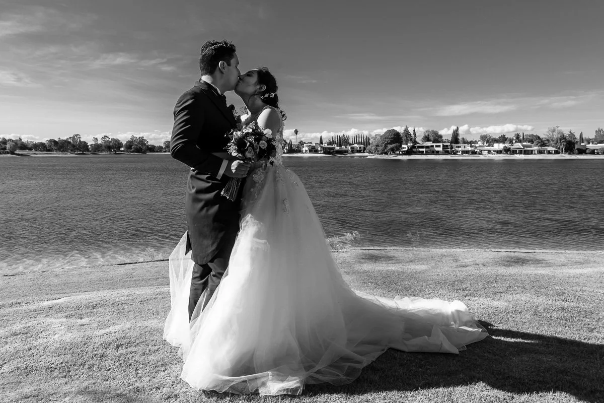 A black and white photo of a newlywed couple kissing by a lake, with the bride in a flowing wedding gown and holding a bouquet of flowers.