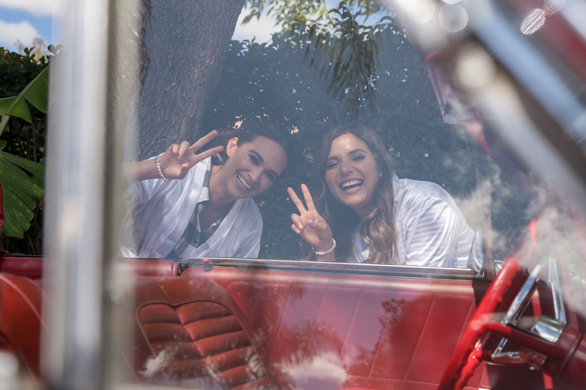 Two smiling women leaning into a car window making peace signs, with a tree and foliage in the background.