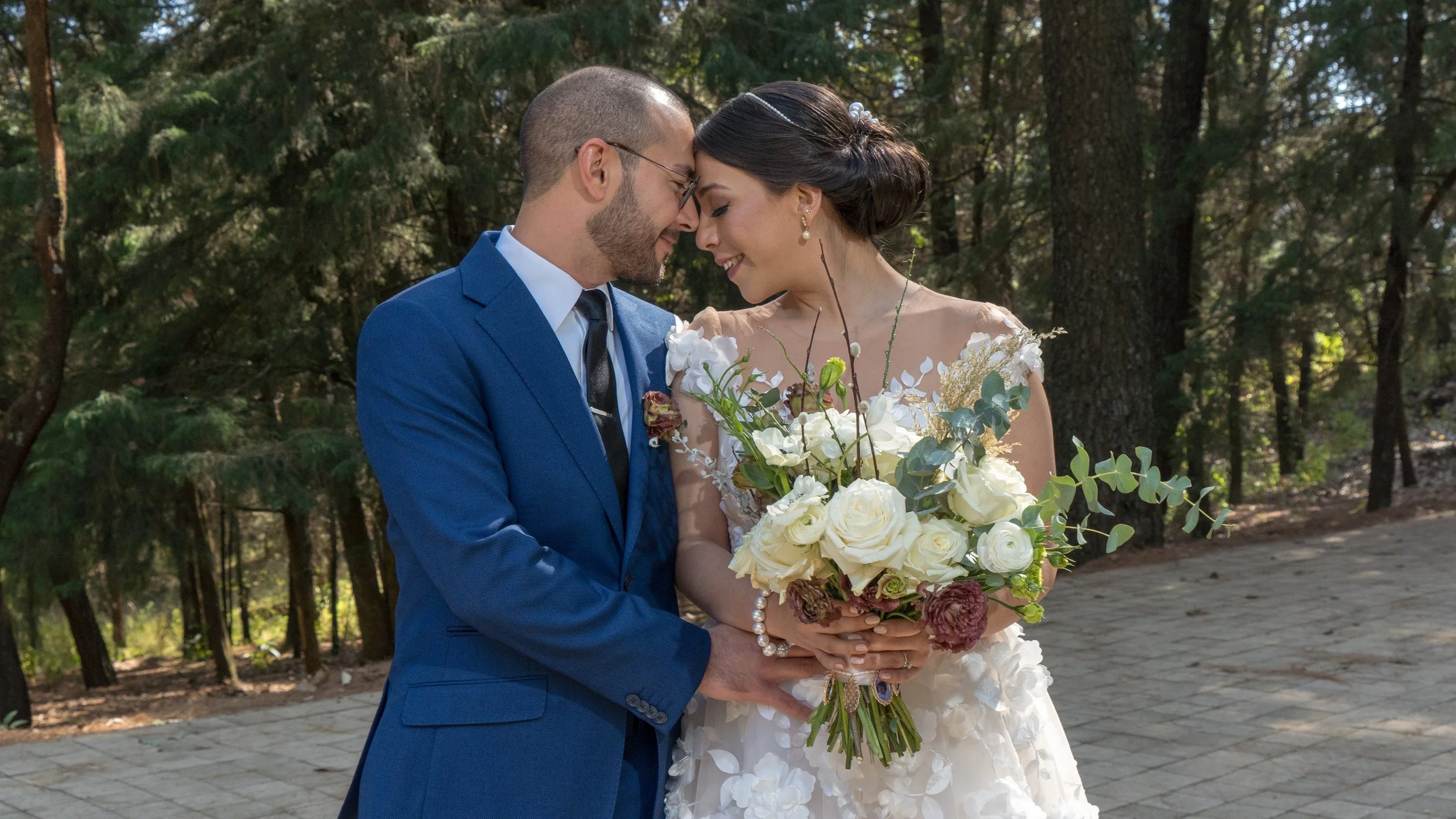 Pareja de novios en boda en el bosque, ambos con las frente tocándose y sosteniéndose las manos. Ella sostiene un ramo de flores blancas y moradas.