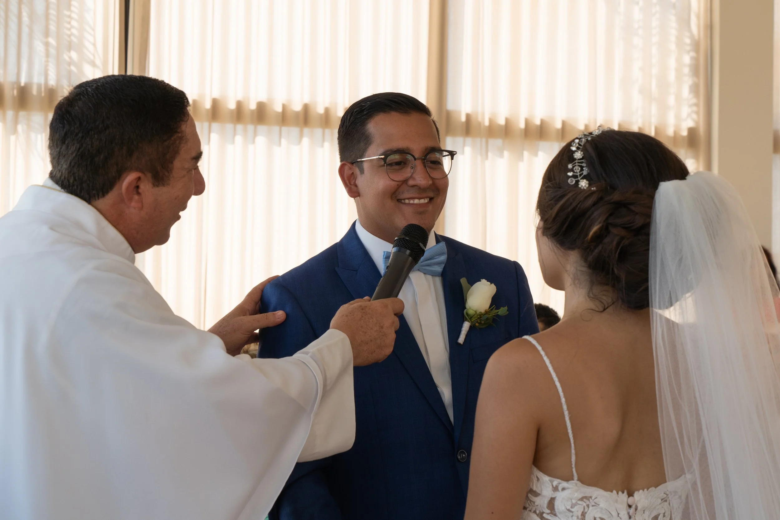 Pareja de novios en ceremonia de boda con un oficiante, en un ambiente interior con cortinas de fondo.