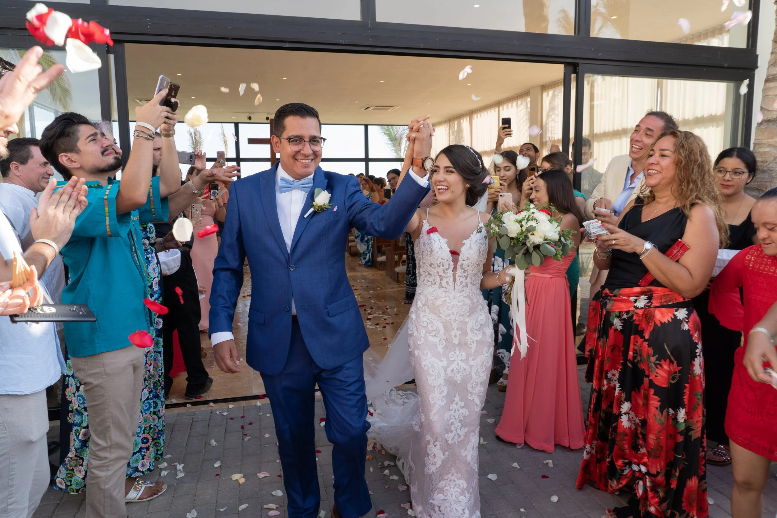 Pareja de recién casados caminando entre amigos y familiares en una celebración de boda, lanzando pétalos de rosa mientras la pareja sonríe y se saludan.