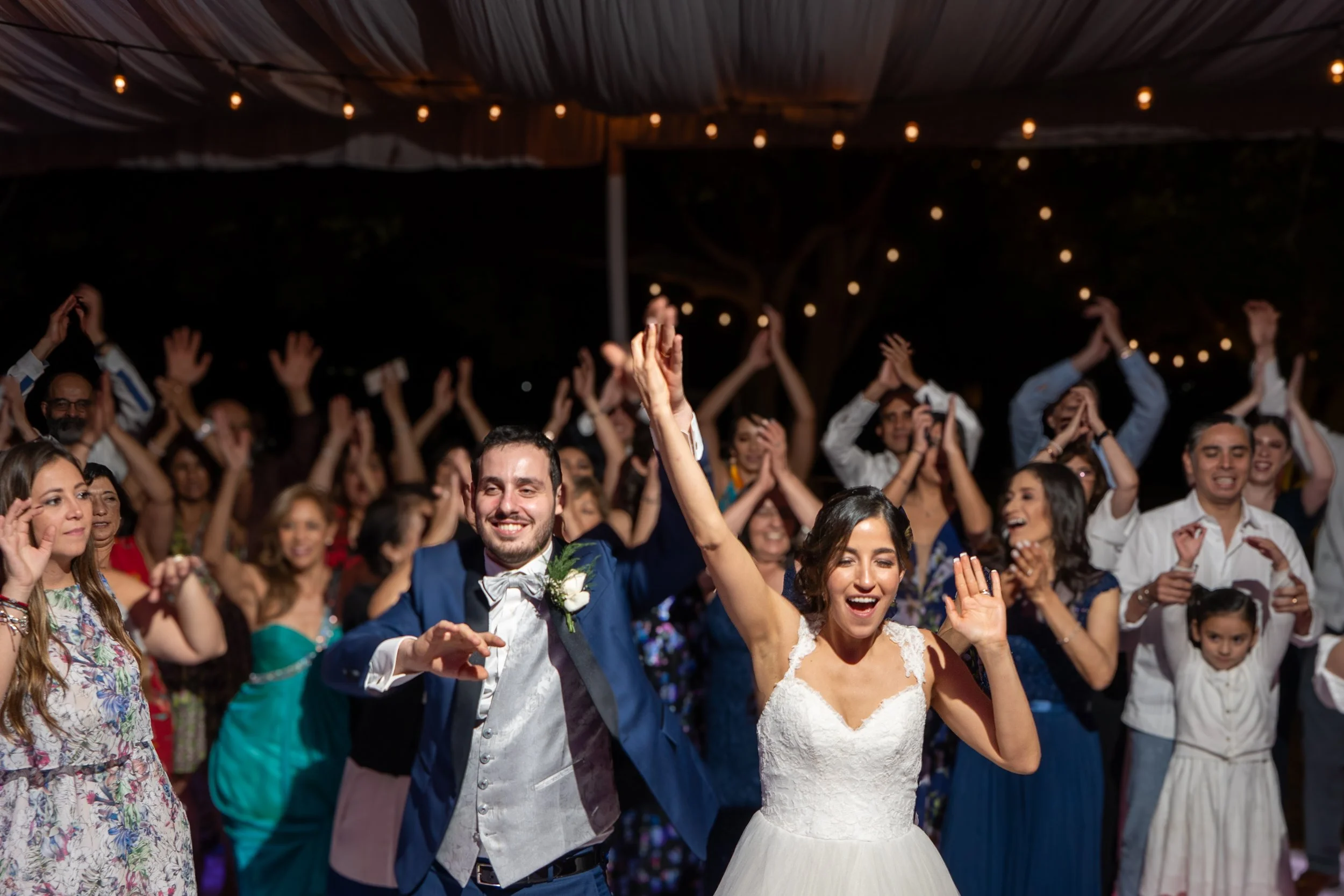 Pareja de recién casados celebrando con sus invitados en una boda. La novia y el novio están sonriendo y levantando las manos. Hay muchas personas en el fondo bailando y disfrutando.