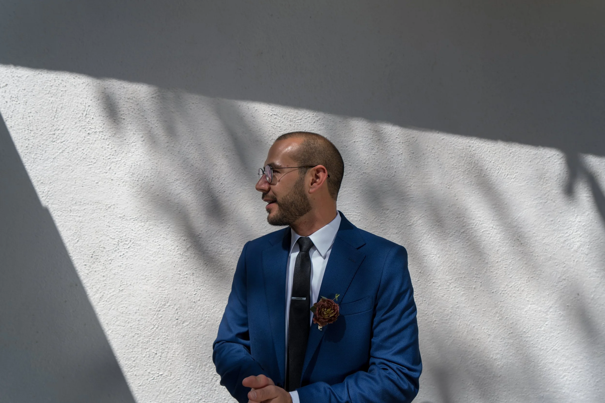Hombre con barba, gafas y traje azul con corbata negra y rosa en la solapa, de perfil, frente a una pared blanca con sombra de ramas y hojas.