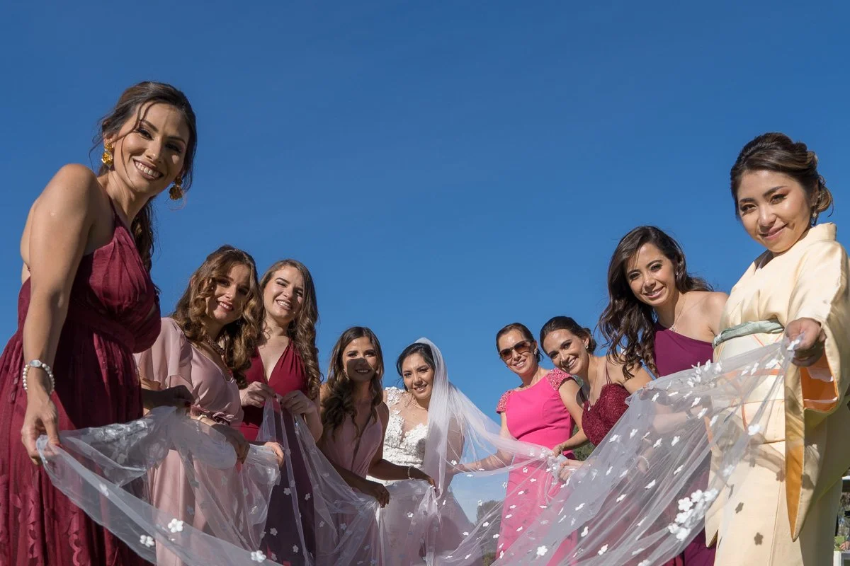 Group of women, including a bride in a wedding dress with a veil, smiling and holding a large, translucent white fabric with floral decorations, outdoors under a clear blue sky.