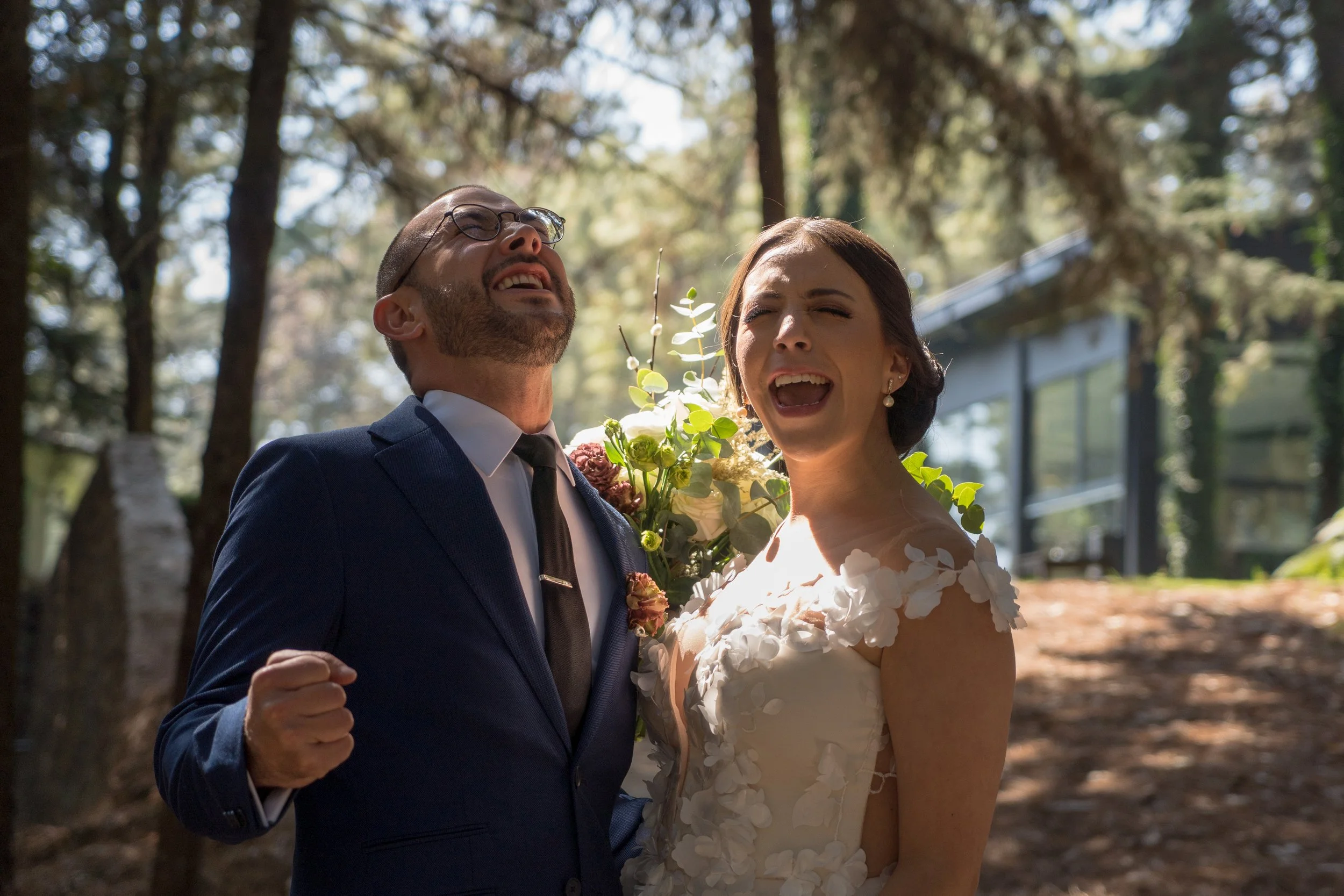 Pareja de recién casados en ceremonia al aire libre en el bosque, ella lleva vestido de novia con adornos florales y él viste traje azul con corbata negra, ambos expresan alegría y emoción.