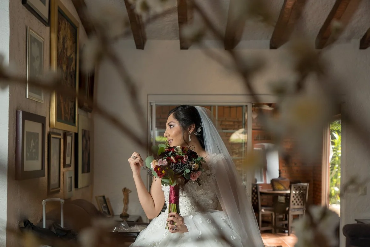 A bride in a white wedding gown and veil holding a bouquet of flowers, standing inside a room with framed artwork on the wall and a wooden ceiling with exposed beams.