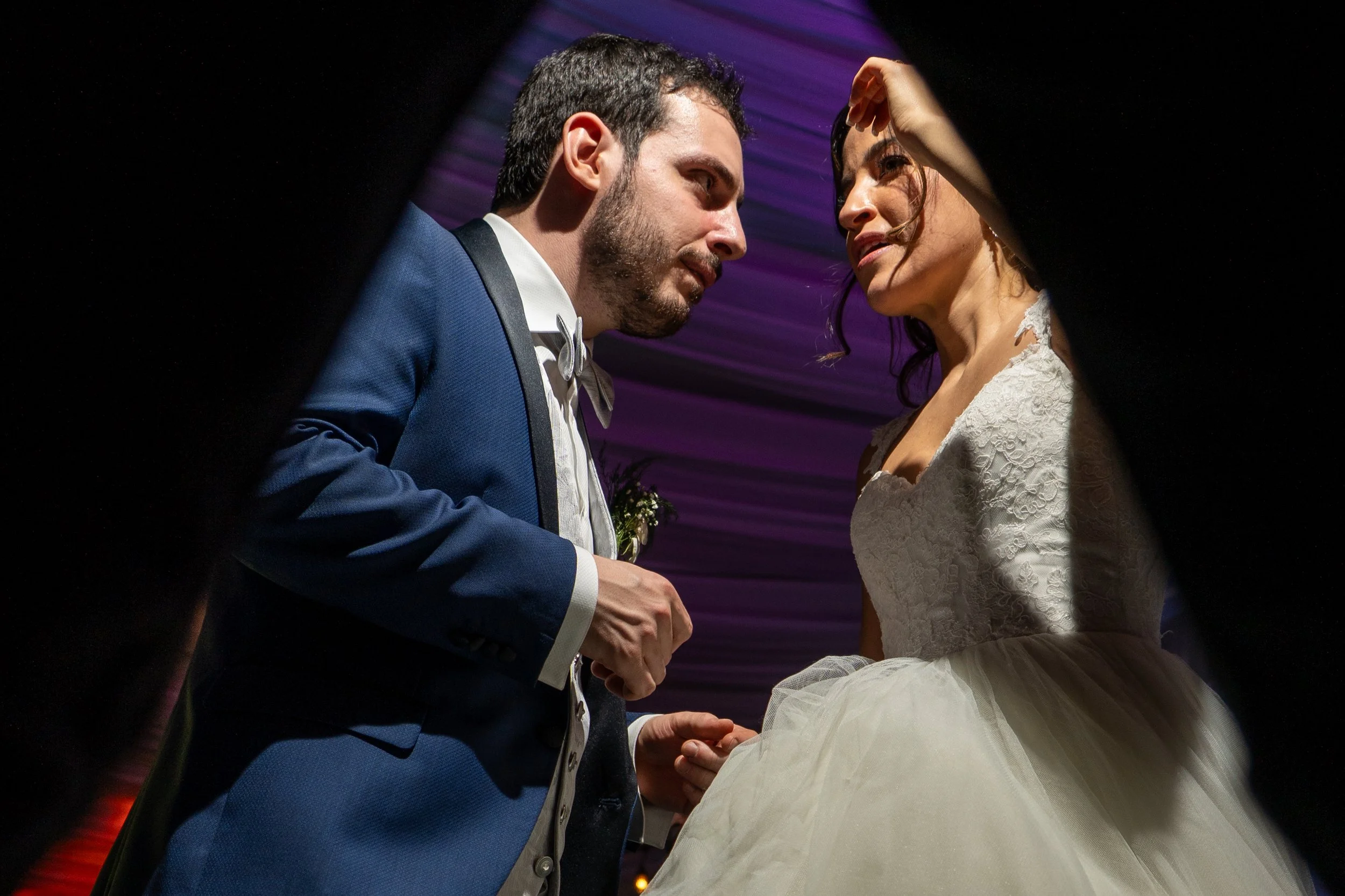 Boda con un hombre vestido con traje azul y una mujer con vestido de novia, en una ceremonia bajo iluminación colorida.