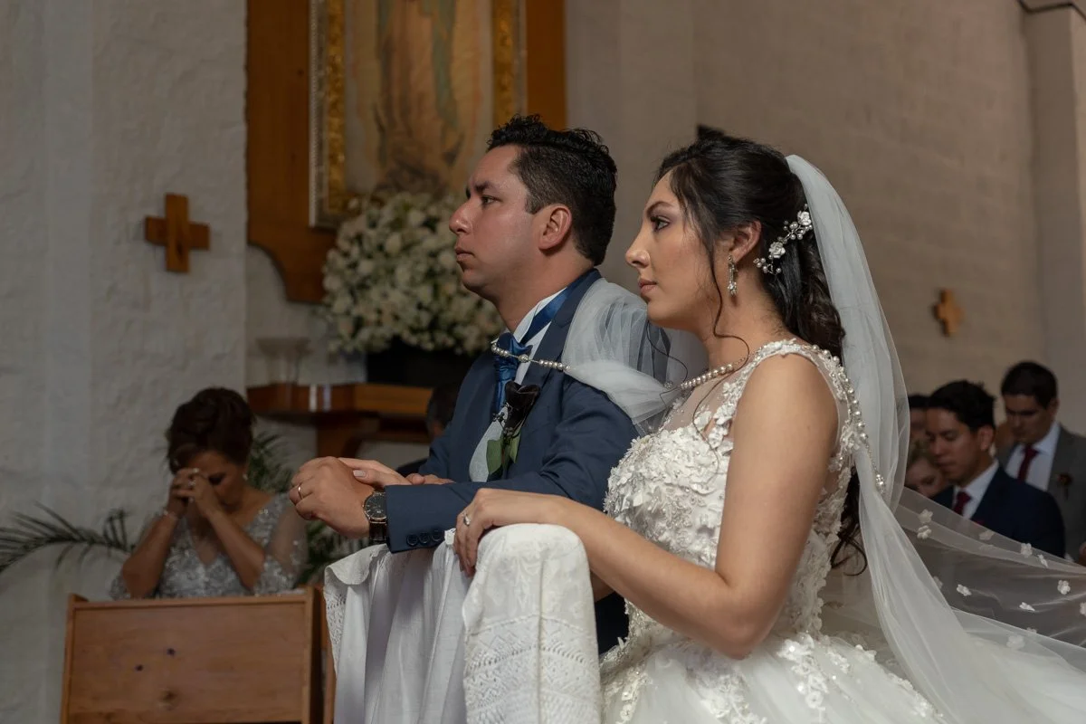 A bride and groom kneel during a wedding ceremony inside a church, with the bride wearing a white wedding gown and veil, and the groom in a blue suit, both looking serious and focused.