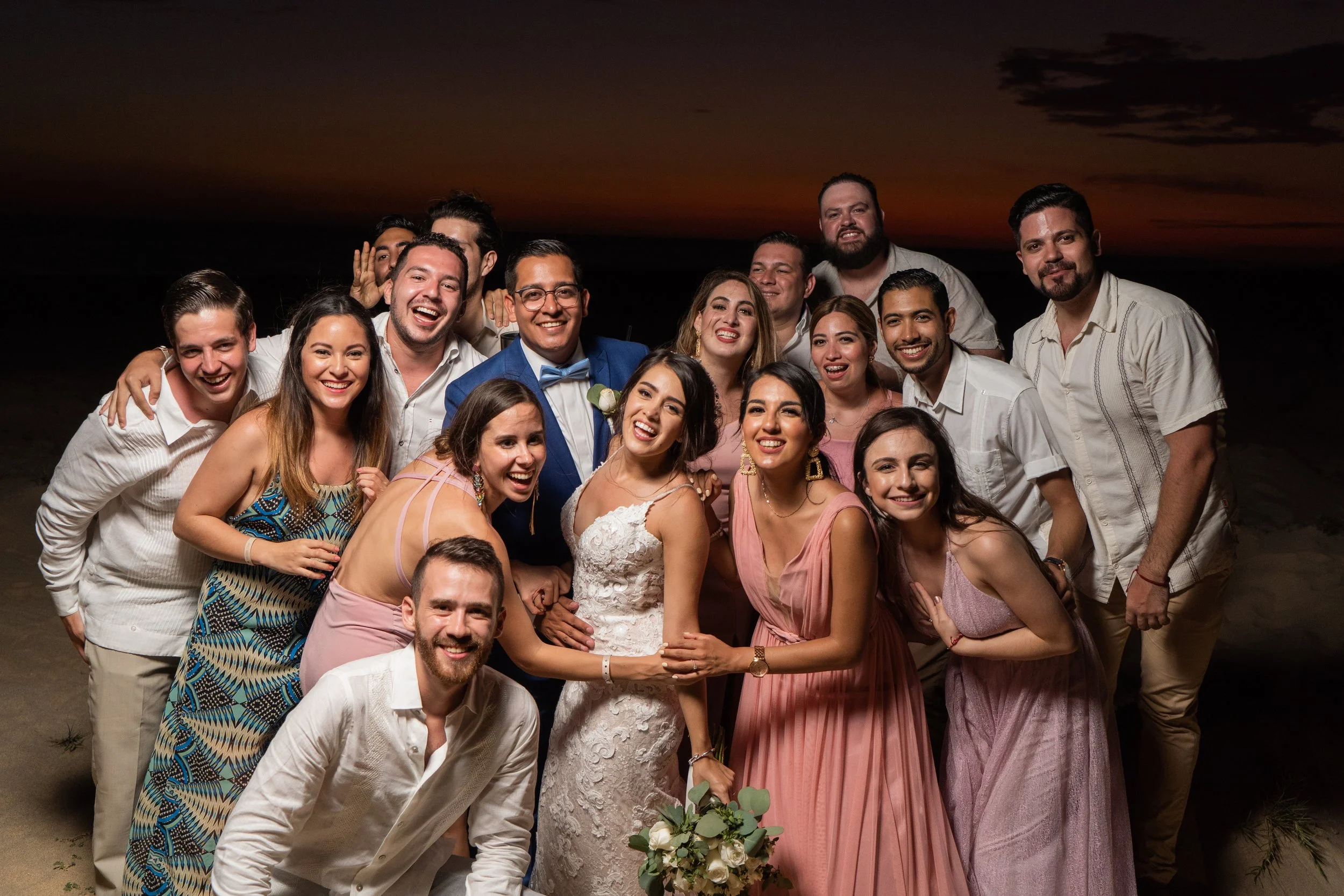 Grupo de personas en una celebración, probablemente una boda, en la playa al atardecer, todas sonriendo y vestidas de manera formal y festiva.