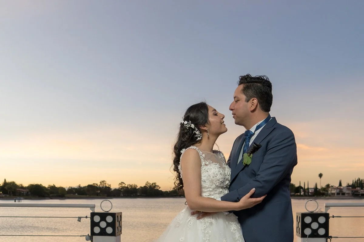 A bride and groom stand close together on a dock at sunset, embracing each other with the water and trees in the background.