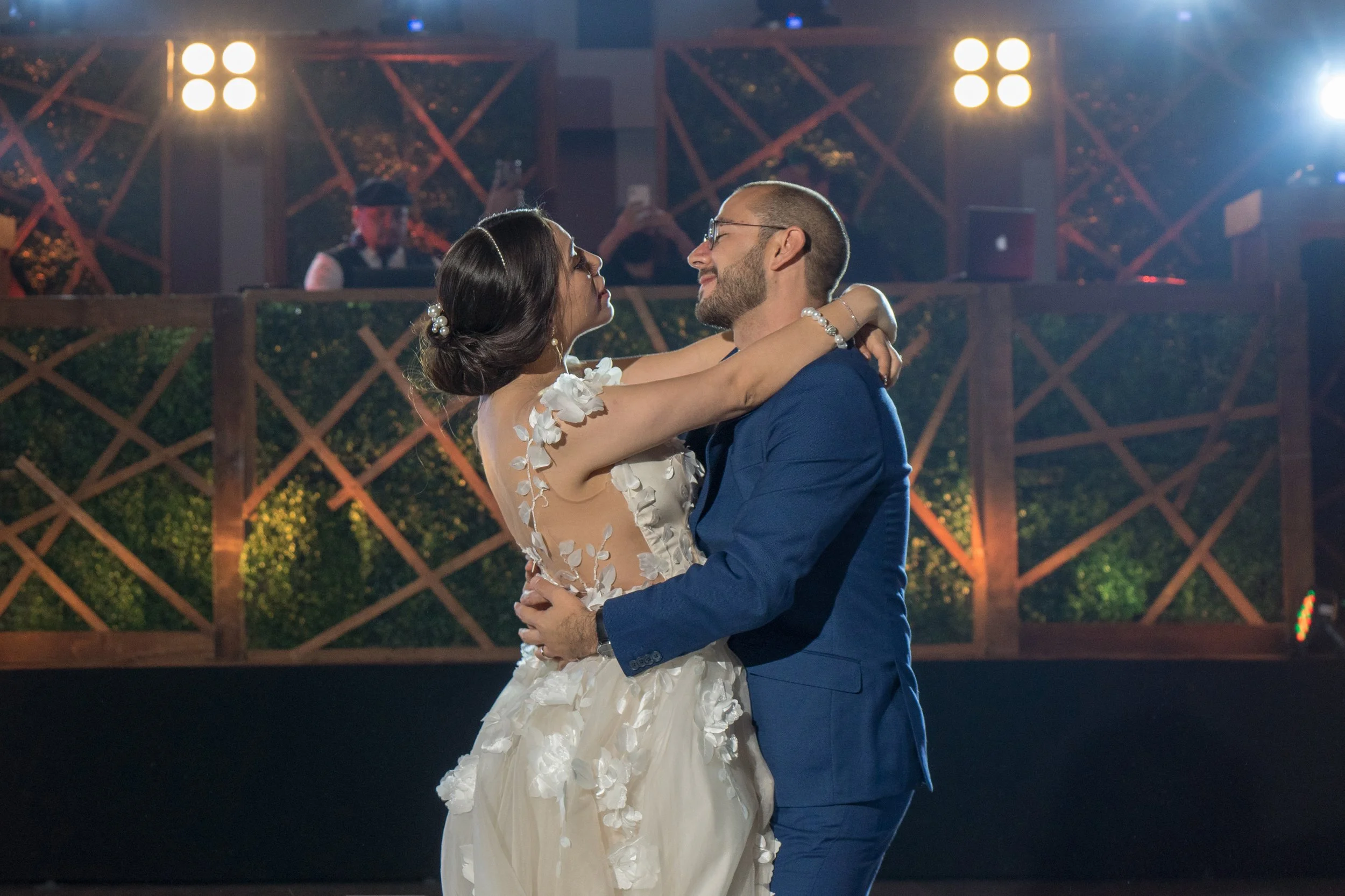 Pareja bailando en una boda, la mujer lleva vestido blanco con detalles florales y el hombre traje azul. Fondo con iluminación y alguien tomando fotos.