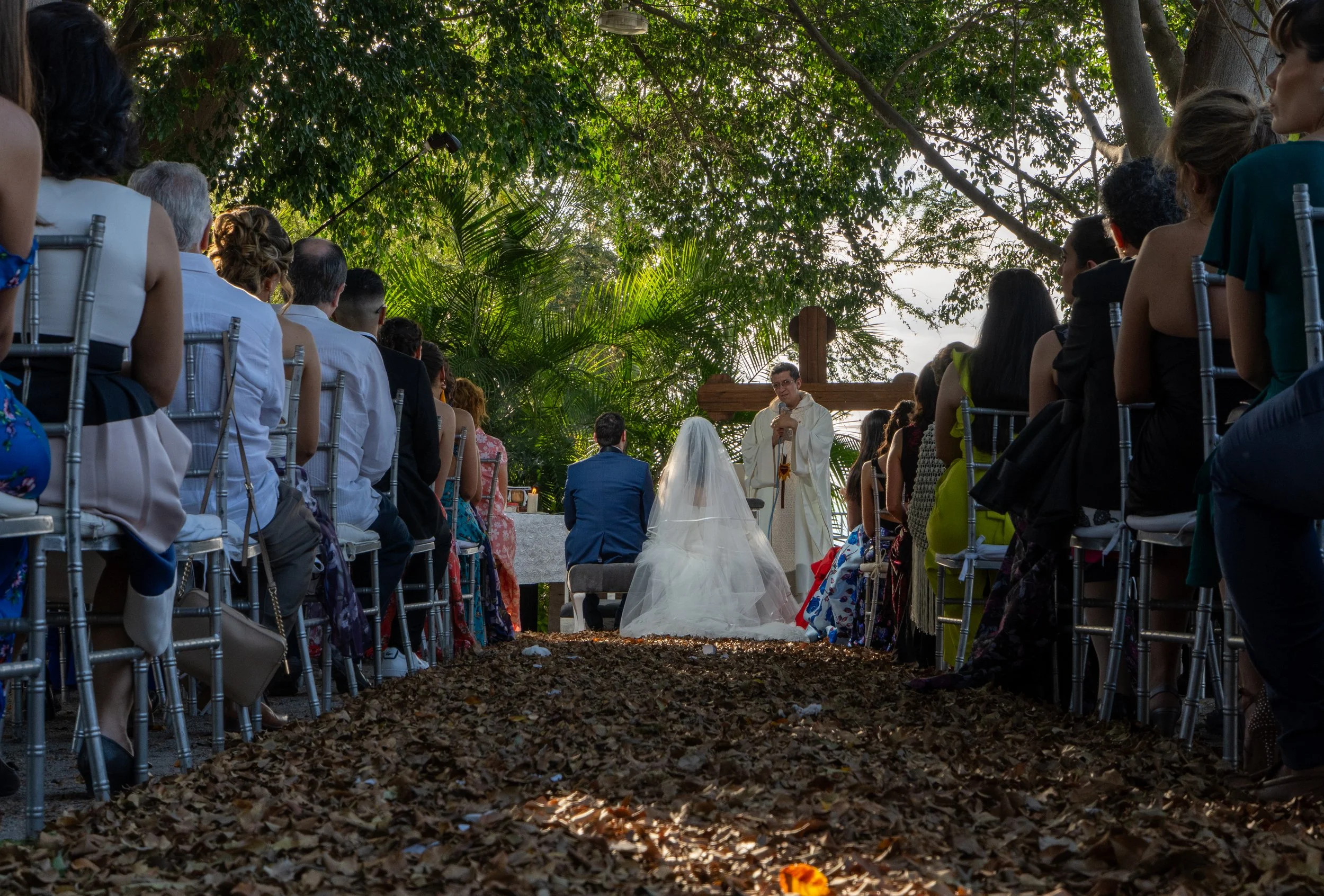 Boda al aire libre con pareja en la ceremonia, asistidos por un oficiante, rodeados de invitados sentados en sillas plateadas, en un entorno natural con árboles y plantas verdes.