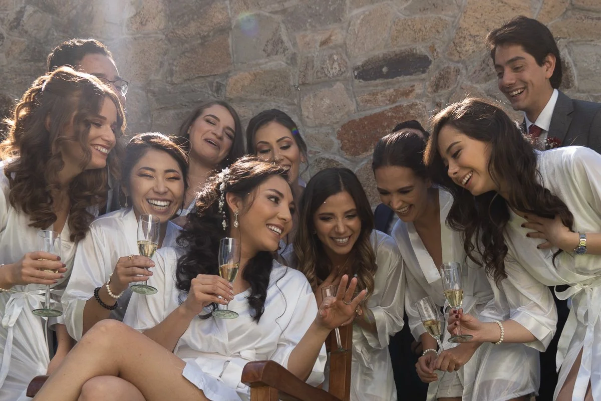 A group of women and one man dressed in robes celebrating, with several holding champagne glasses, in a room with a stone wall background.