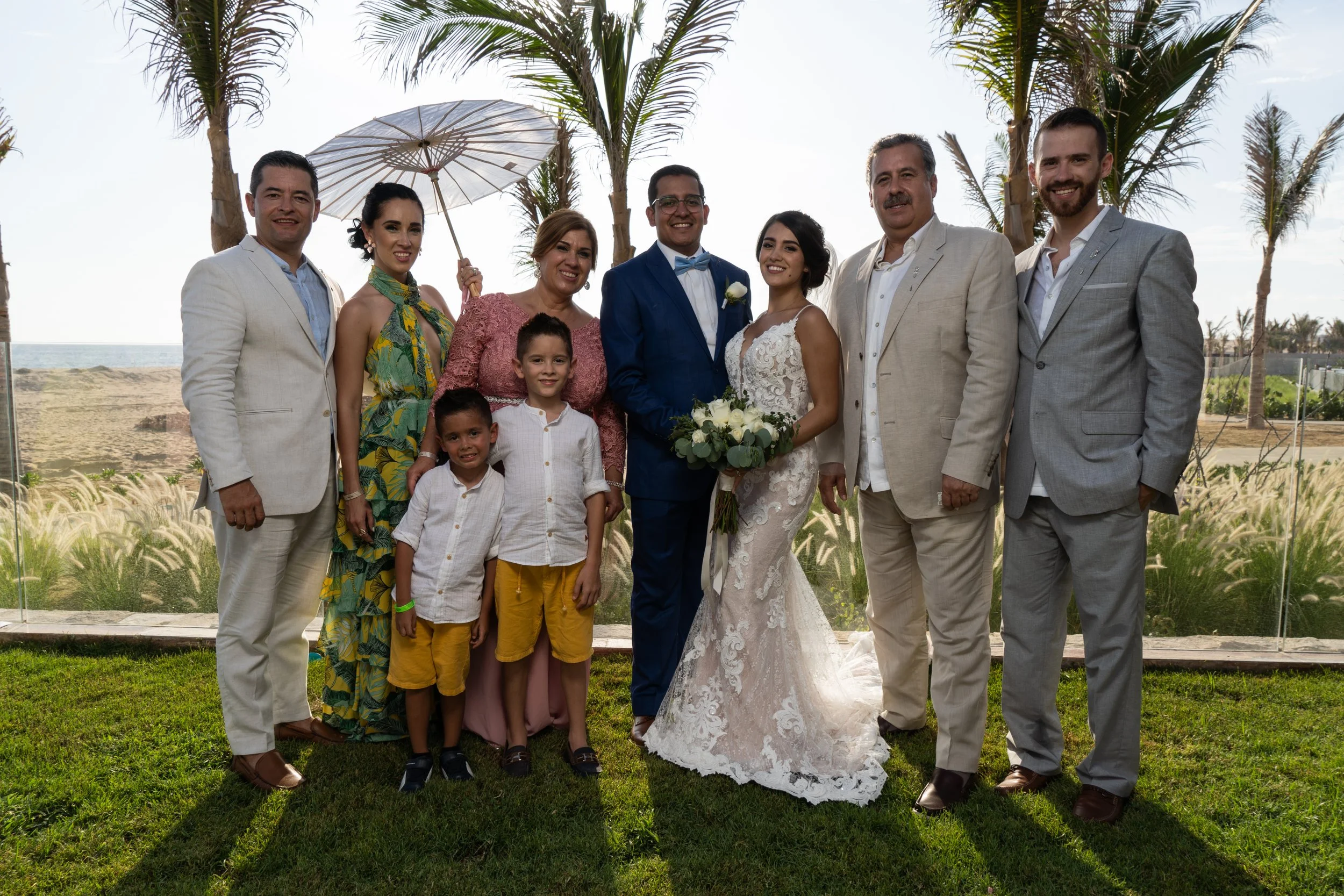 Grupo de personas en una boda al aire libre con palmeras y vista al mar de fondo, incluyendo a la novia con vestido de encaje y ramo de flores, y al novio con traje azul, rodeados de familiares y amigos en atuendos formales y casuales, algunos niños 