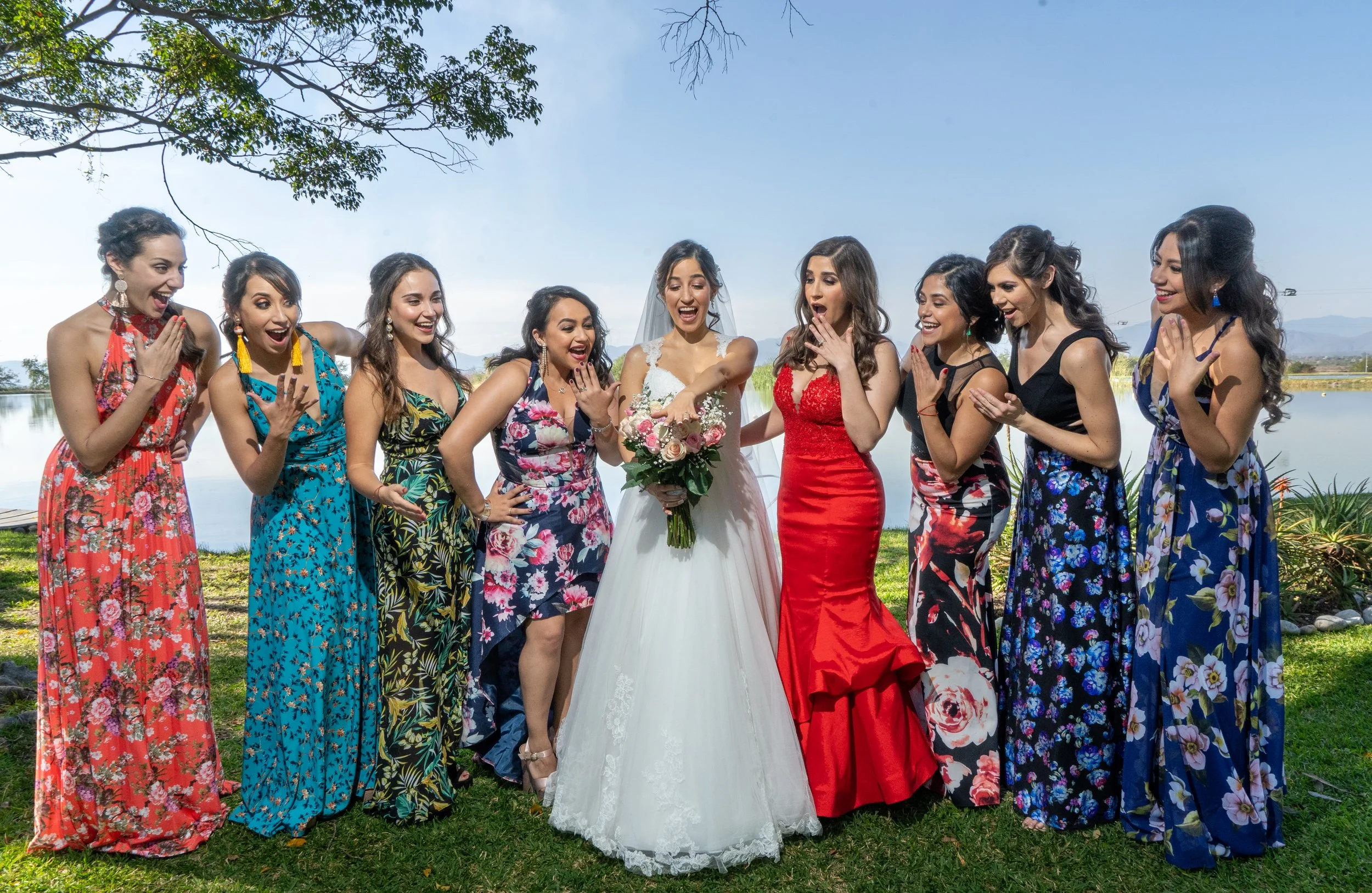 Grupo de mujeres en un evento al aire libre, algunas vestidas con vestidos largos coloridos y una mujer en vestido de novia con ramo de flores, todas parecen sorprendidas y felices.