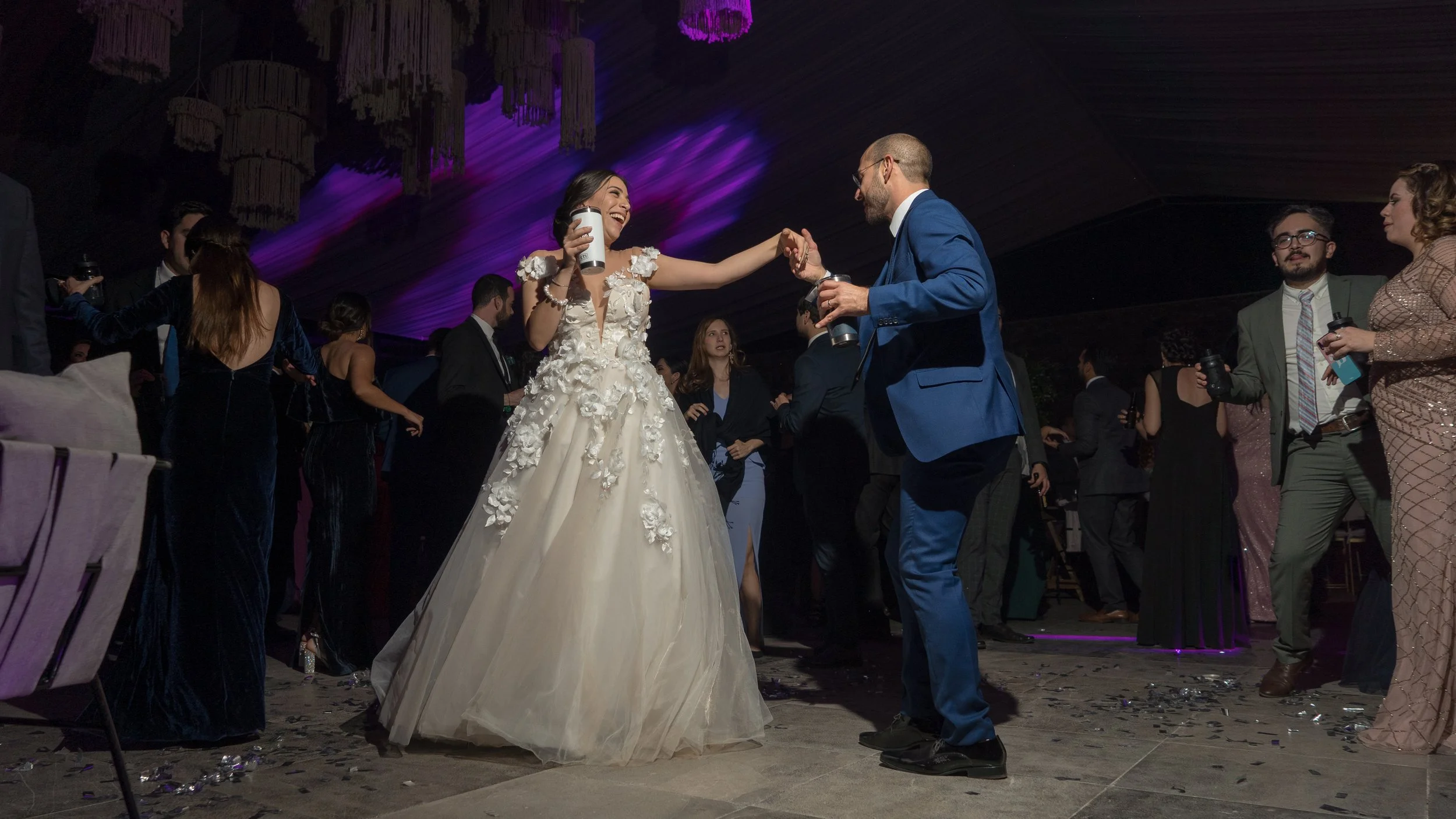 Pareja abrazándose y sonriendo en la pista de baile de una fiesta de boda con invitados bailando y tomando fotografías.