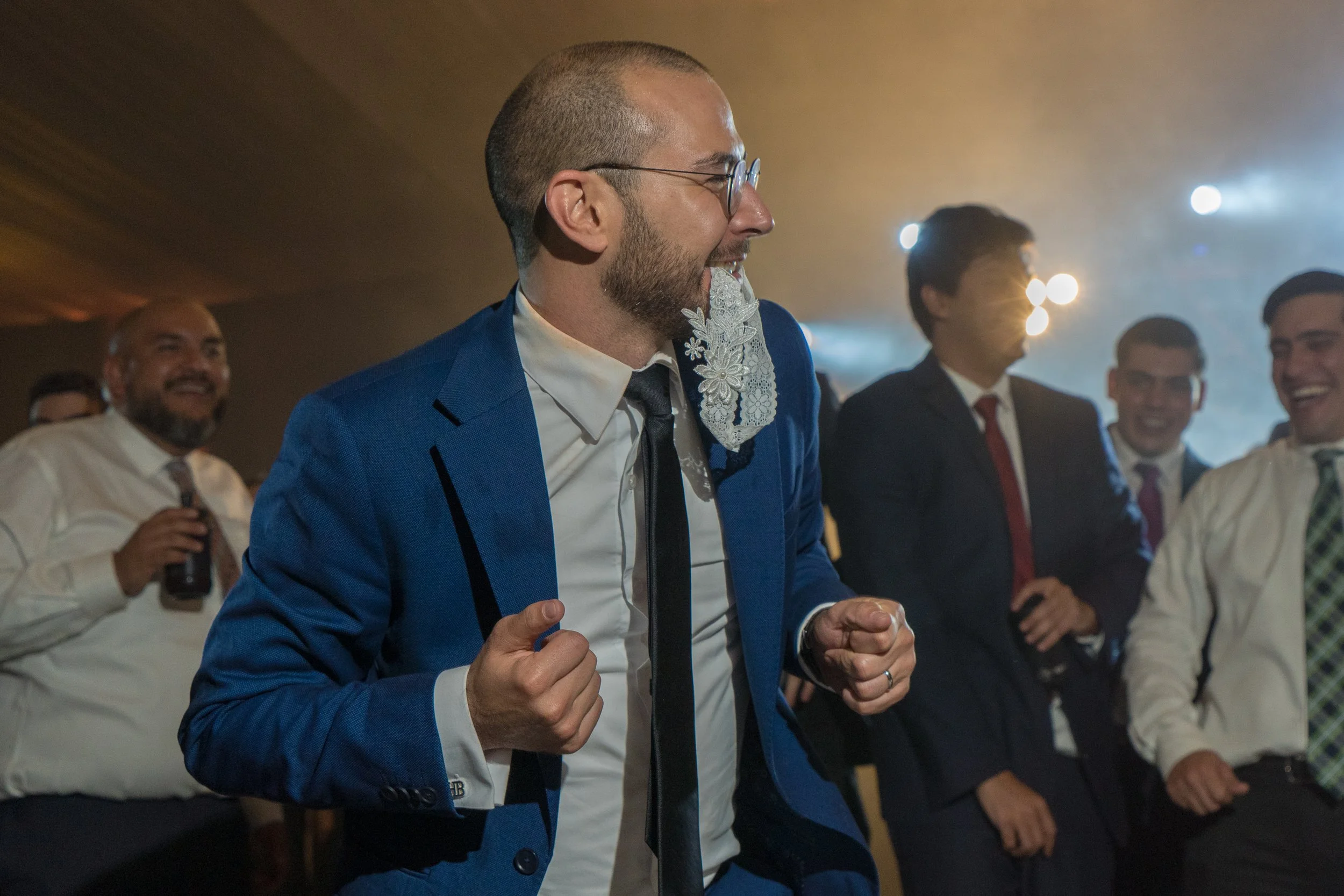 Hombre con barba, gafas, traje azul y corbata negra, celebrando con entusiasmo en un evento, rodeado de varias personas igualmente vestidas formalmente, en un ambiente de celebración con luces brillantes.