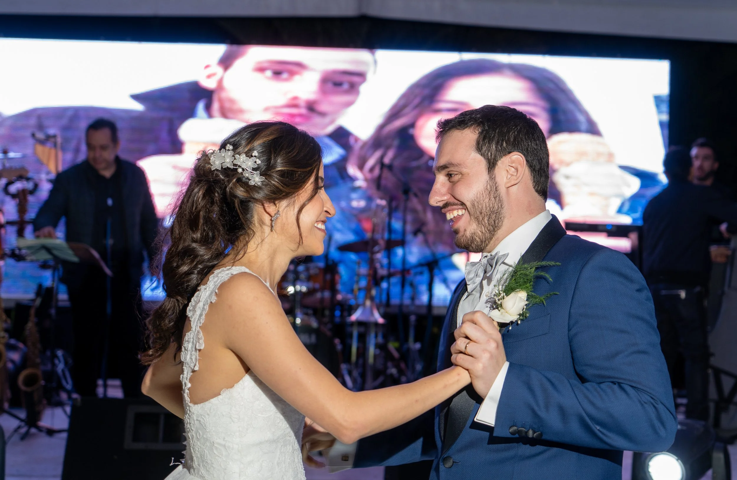 Una pareja de novios bailando en su boda, ella con vestido blanco y él con traje azul, sonriendo y mirándose con felicidad, en un evento con iluminación y una pantalla grande en el fondo.