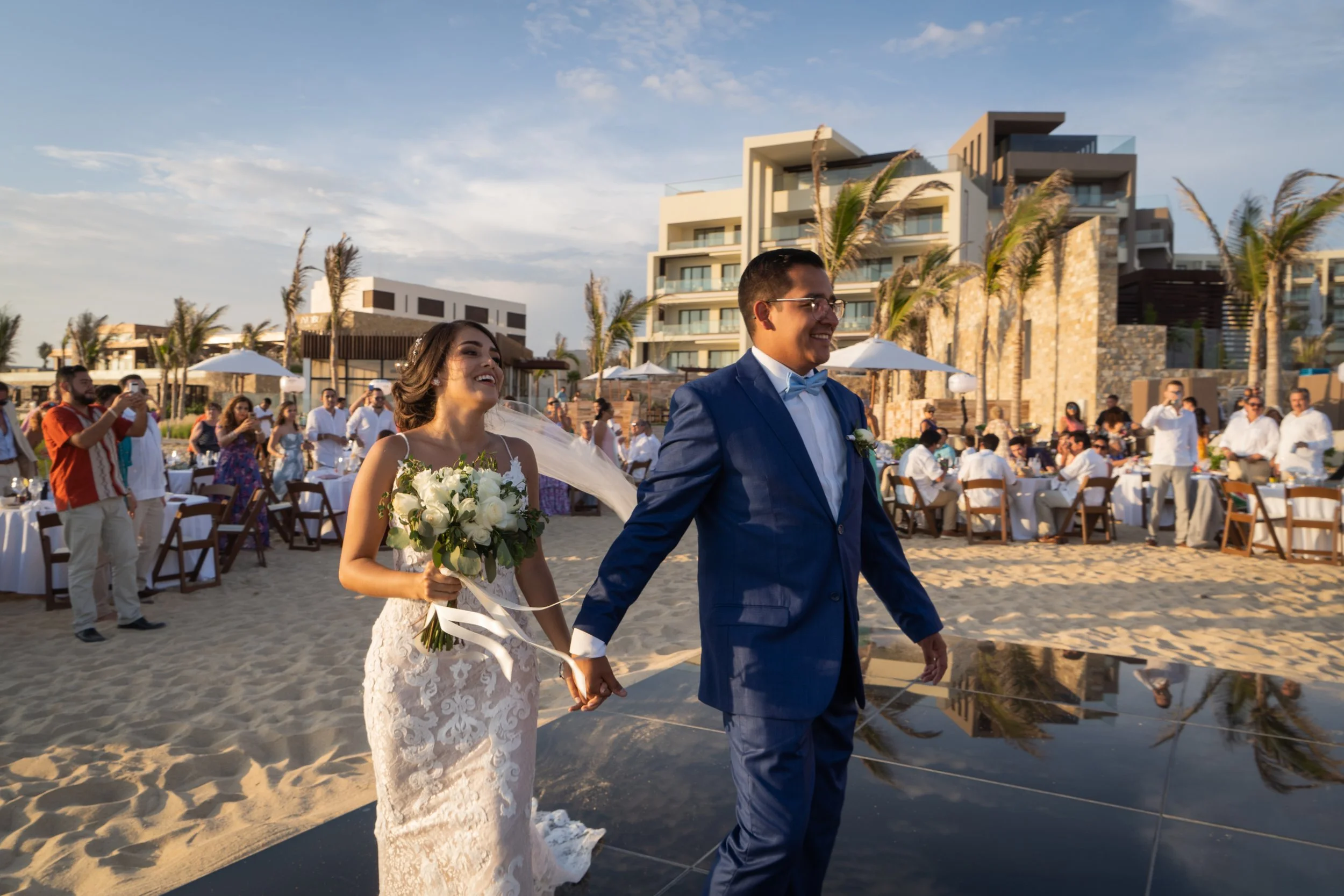 Pareja de recién casados en una boda en la playa, la novia con vestido de encaje y ramo de flores blancas, el novio con traje azul y gafas, rodeados de invitados y edificios modernos en el fondo.