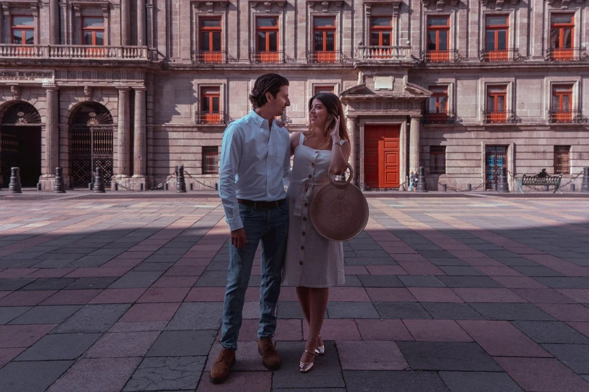 A man and woman standing and looking at each other in front of a historic building with large windows and a red door.