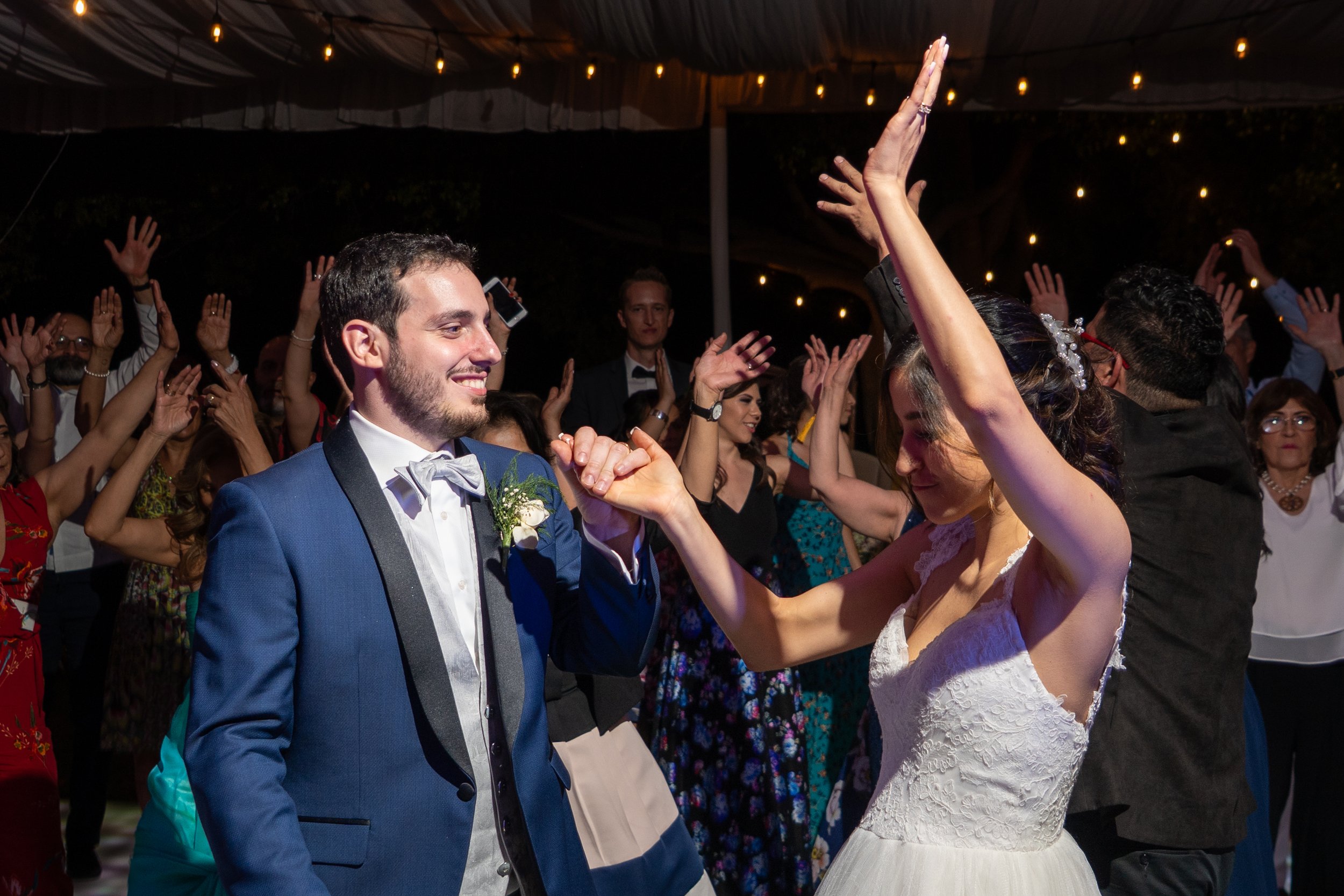 Pareja de recién casados bailando en una fiesta, rodeados de invitados. La pareja está sonriendo y se Observa que la mujer lleva vestido blanco de novia y el hombre traje azul con pajarita. Todos parecen felices en la celebración.
