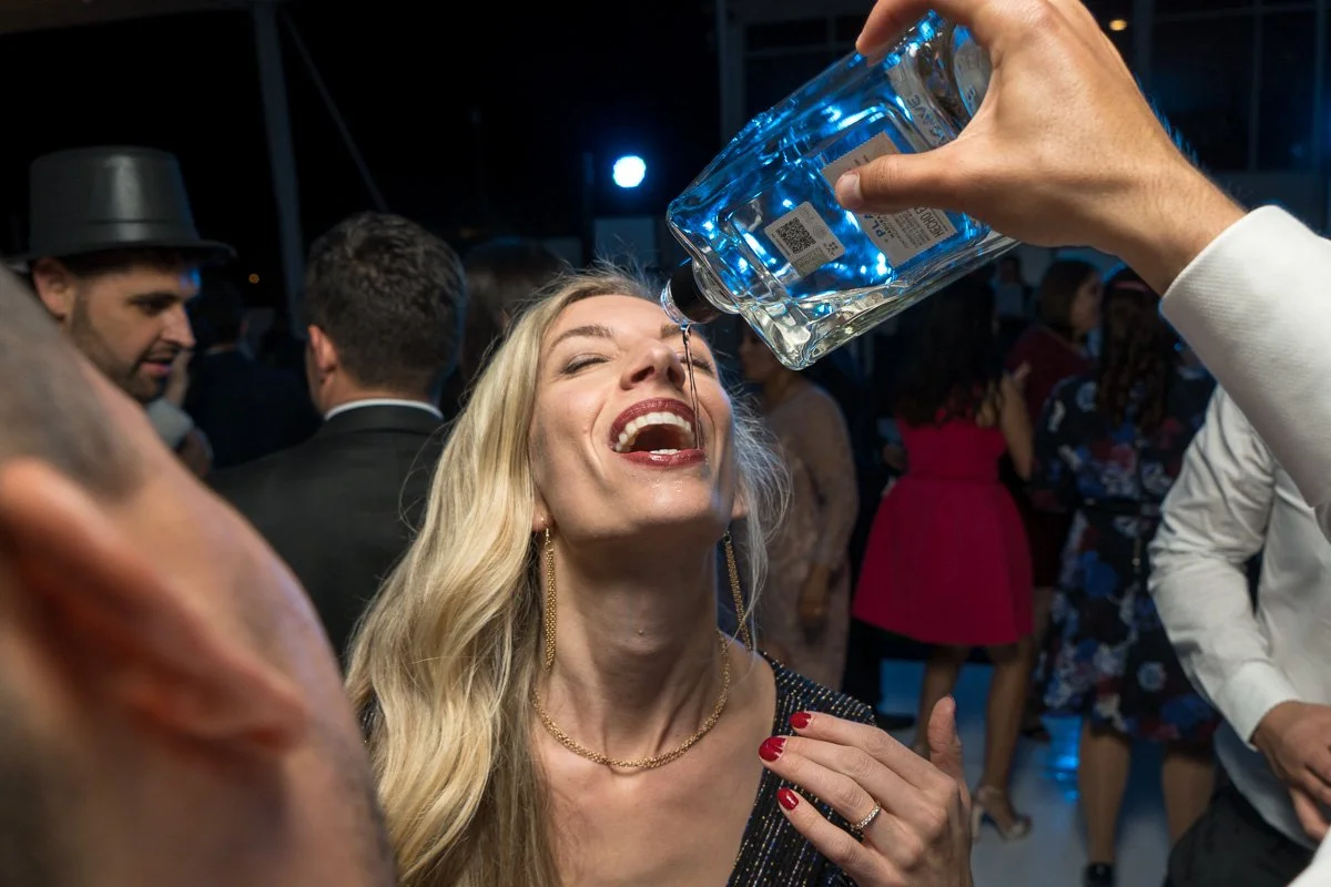 A woman with long blonde hair and red nail polish is smiling and tilting her head back as water from a bottle is poured into her mouth at a party or celebration event.