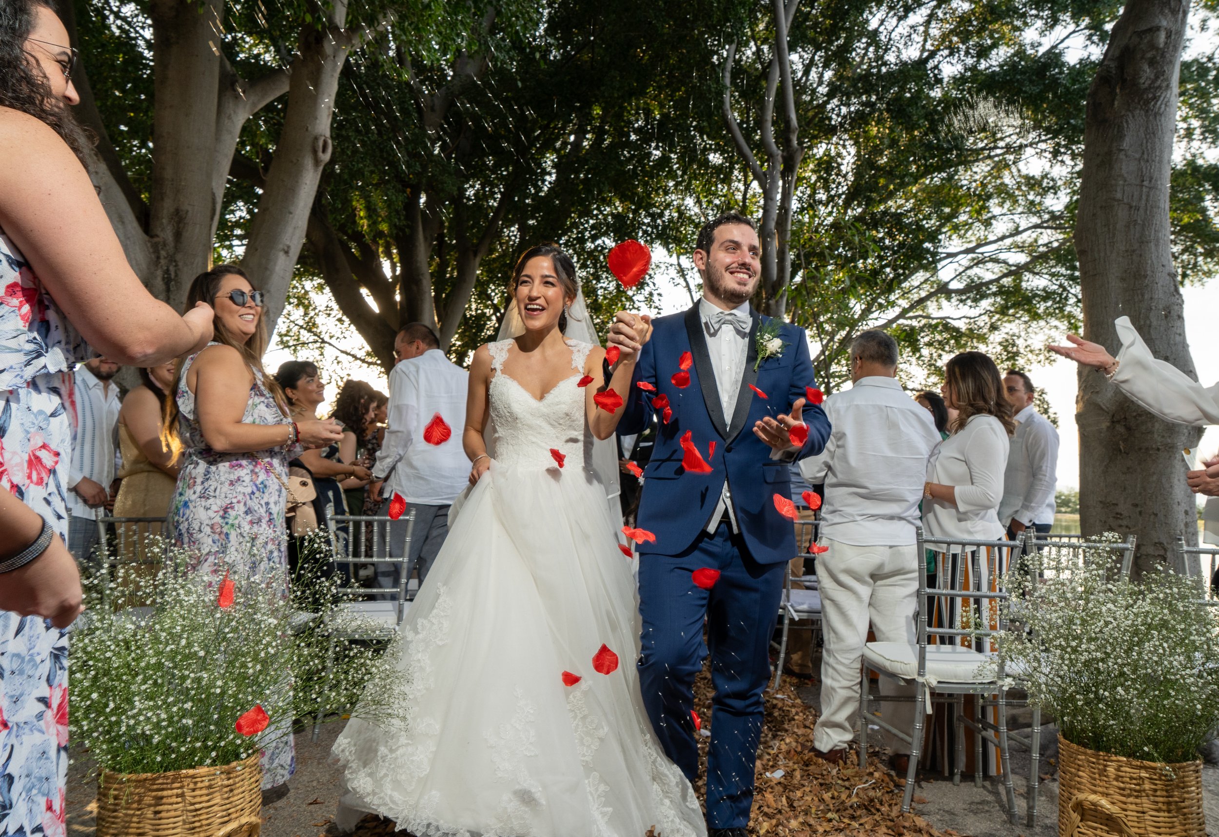 Boda con pareja feliz caminando entre invitados, cayendo pétalos de rosa en un ambiente al aire libre con árboles y decoraciones florales.