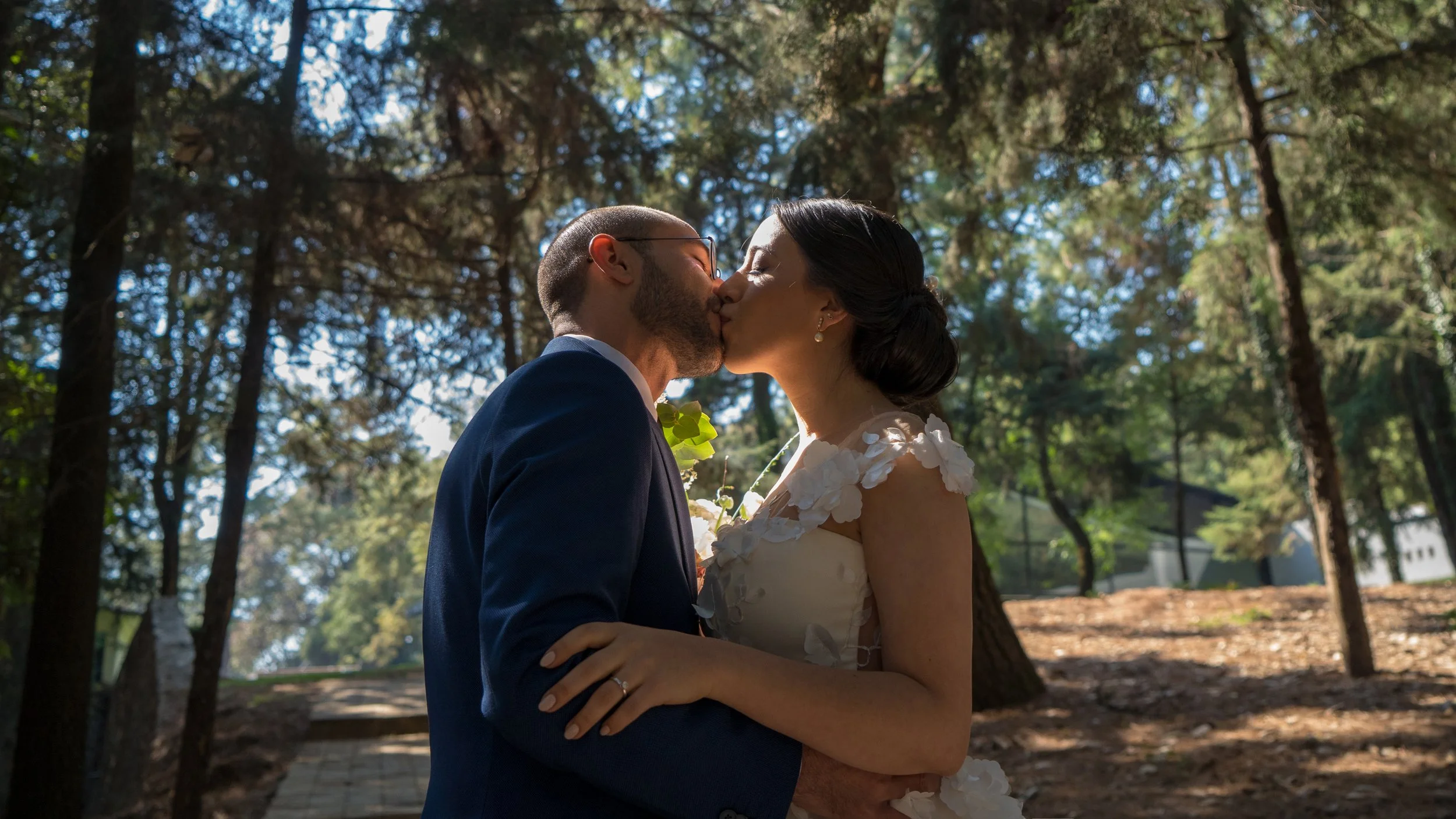 Una pareja se besa en un entorno boscoso, la mujer lleva un vestido de novia y el hombre un traje azul, bajo la sombra de árboles con luz solar filtrándose.