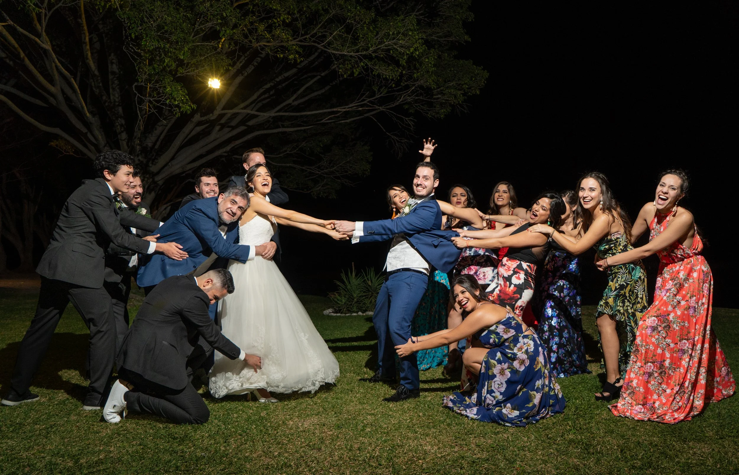 Grupo de personas en una boda, divirtiéndose y haciendo una cadena con la novia y el novio en el centro, en un entorno al aire libre de noche.