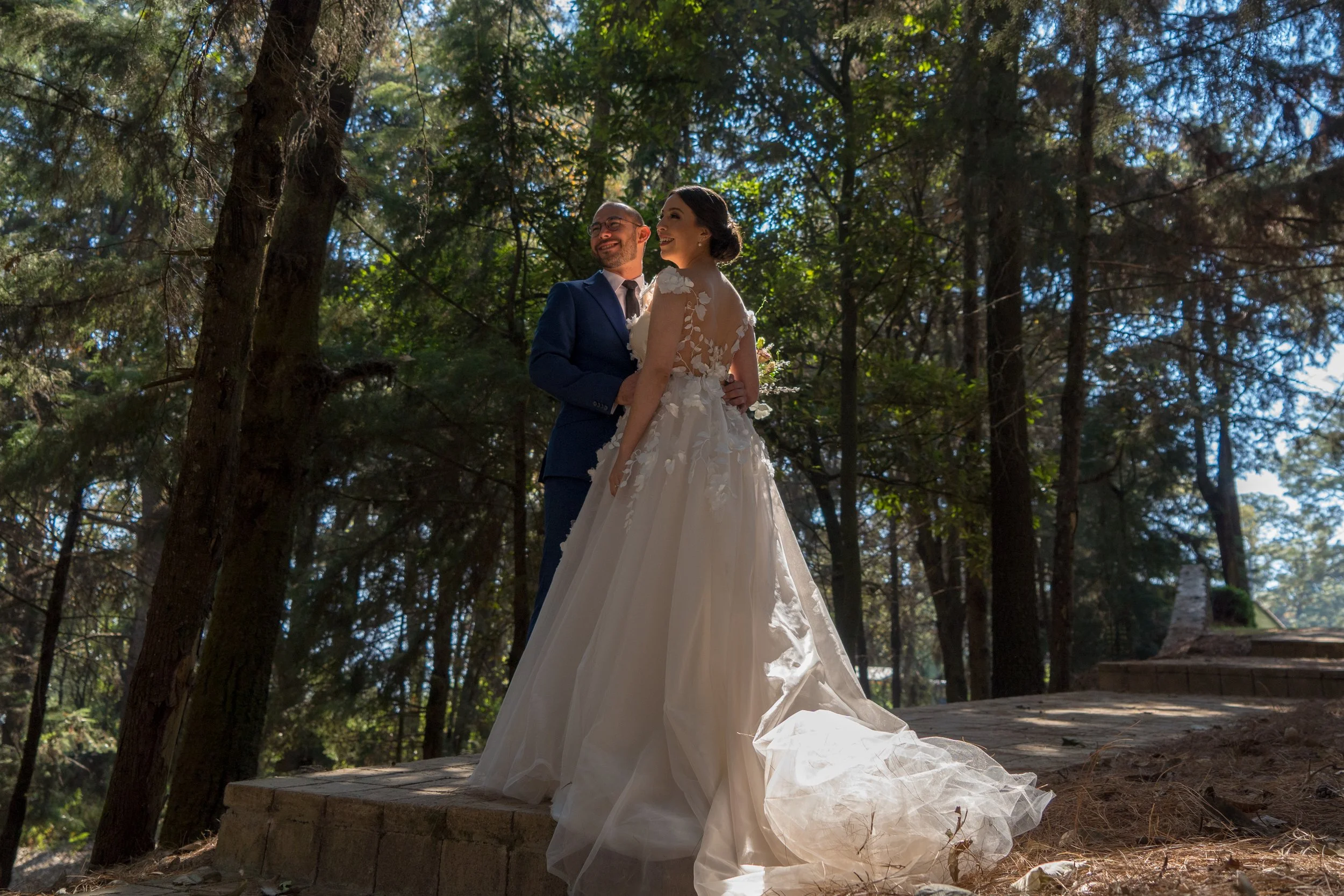 Pareja de recién casados en un bosque, ella con vestido de novia y él con traje, sonriendo y mirándose felizmente.