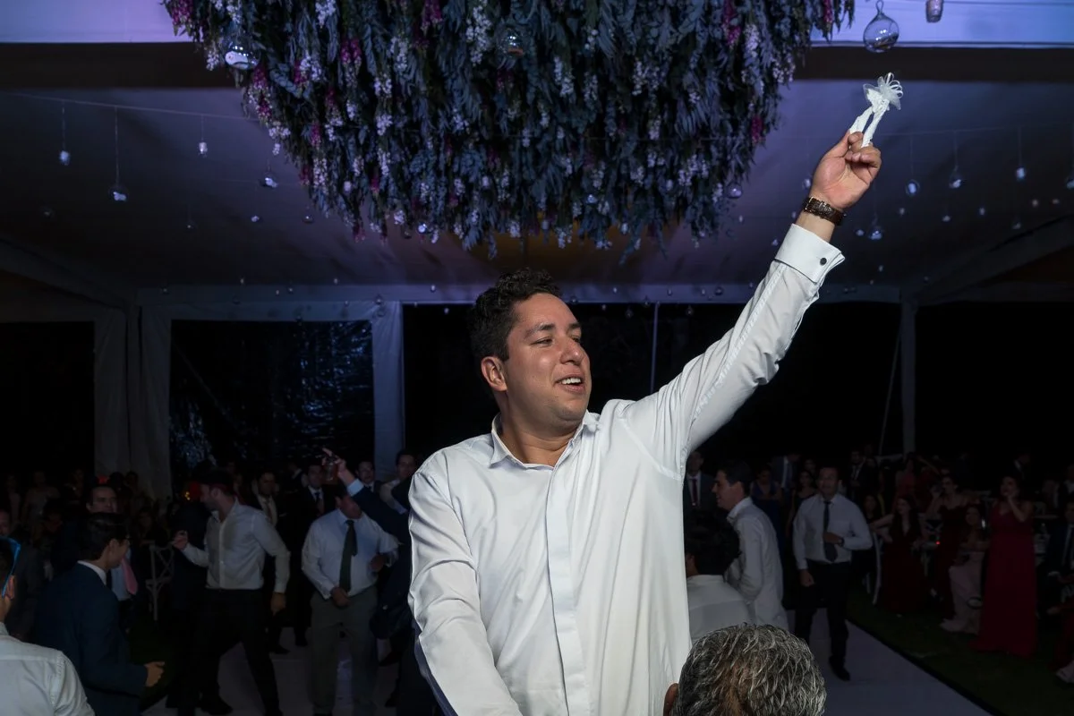 A man in a white shirt dancing at a crowded event, holding a white ribbon and smiling, with a large floral chandelier overhead.
