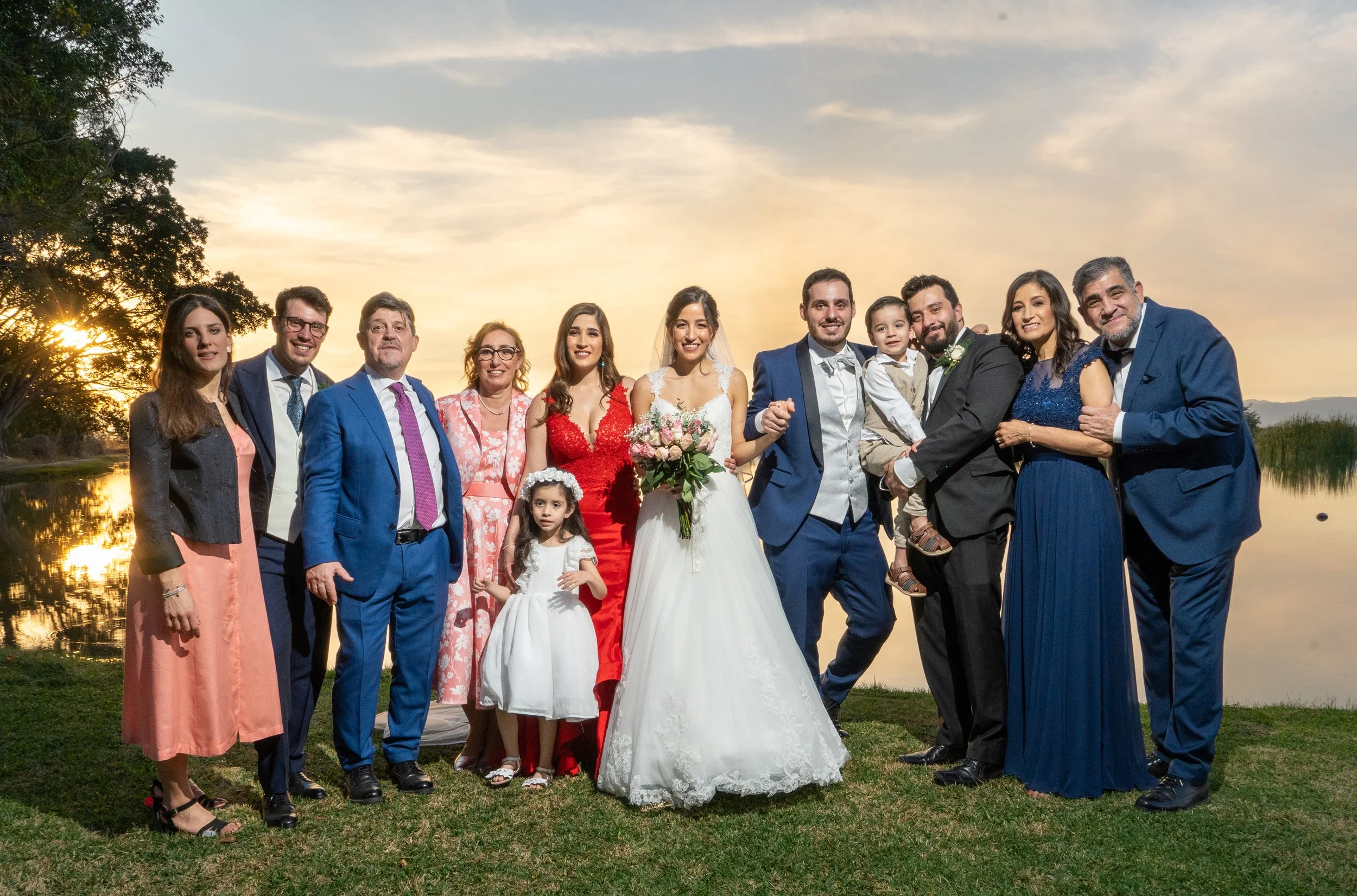 Grupo de personas en celebración de boda junto a un lago durante el atardecer, con la novia en vestido blanco y flores, y todos sonriendo.