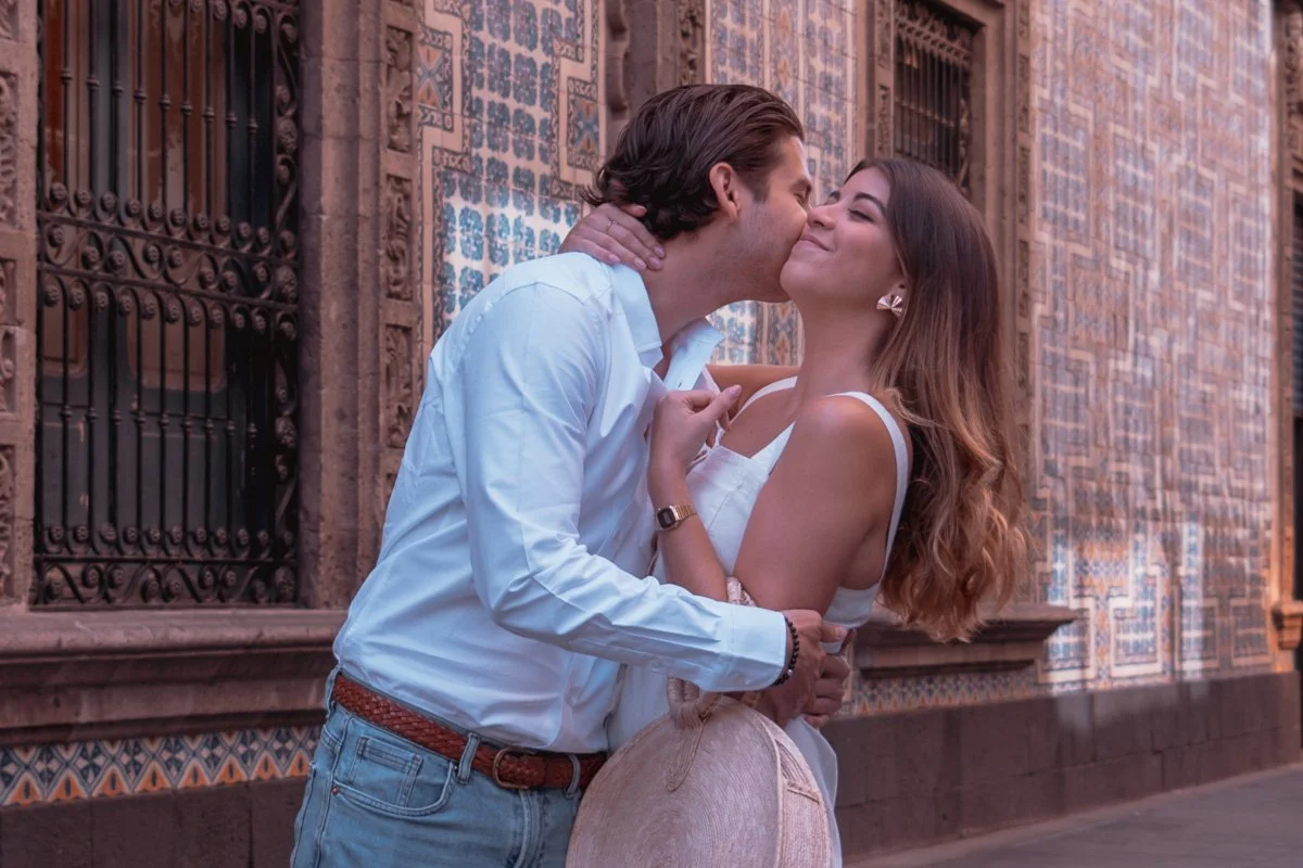 A man and a woman share a romantic moment as he kisses her cheek outside a decorative tiled building.