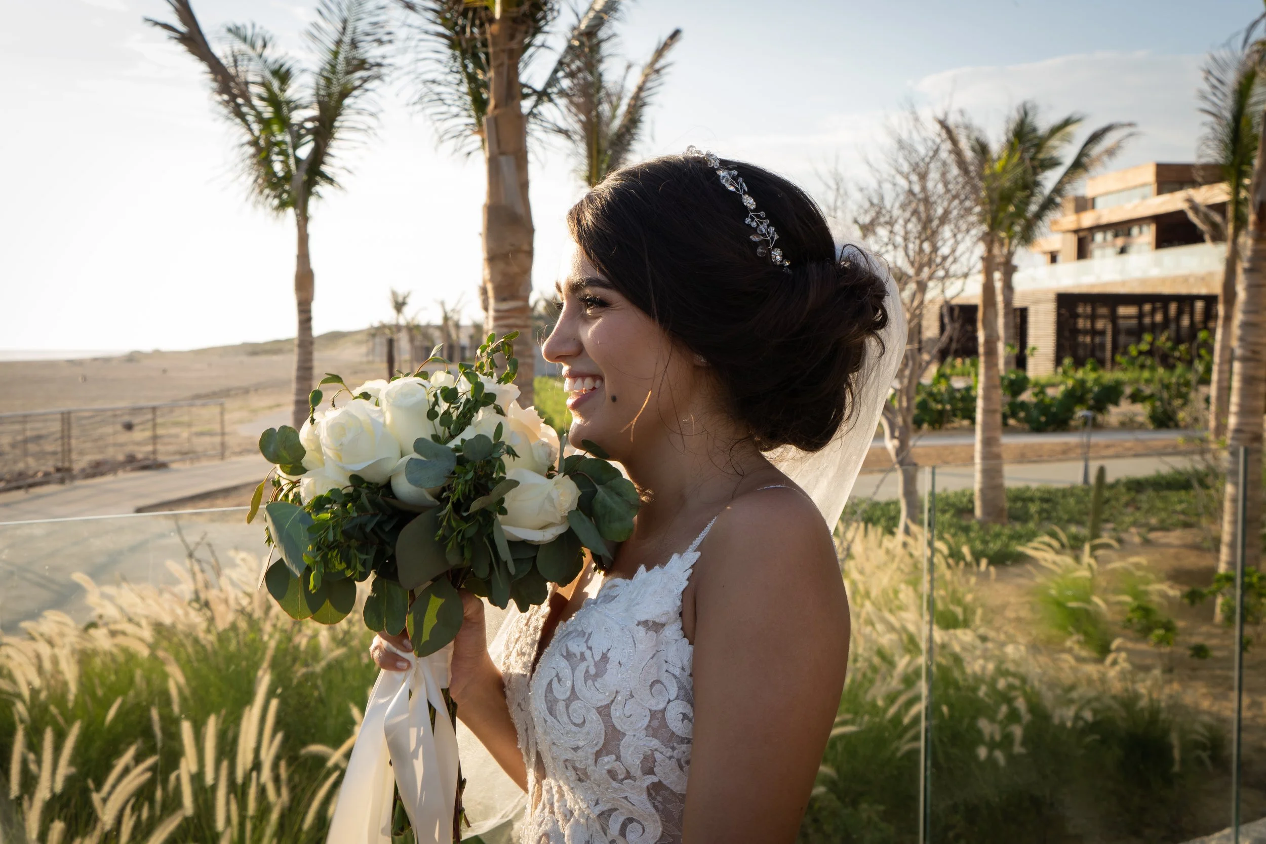 Mujer en vestido de novia sosteniendo ramo de flores blancas y verdes, en un entorno al aire libre con palmeras y edificios de fondo, durante el atardecer.